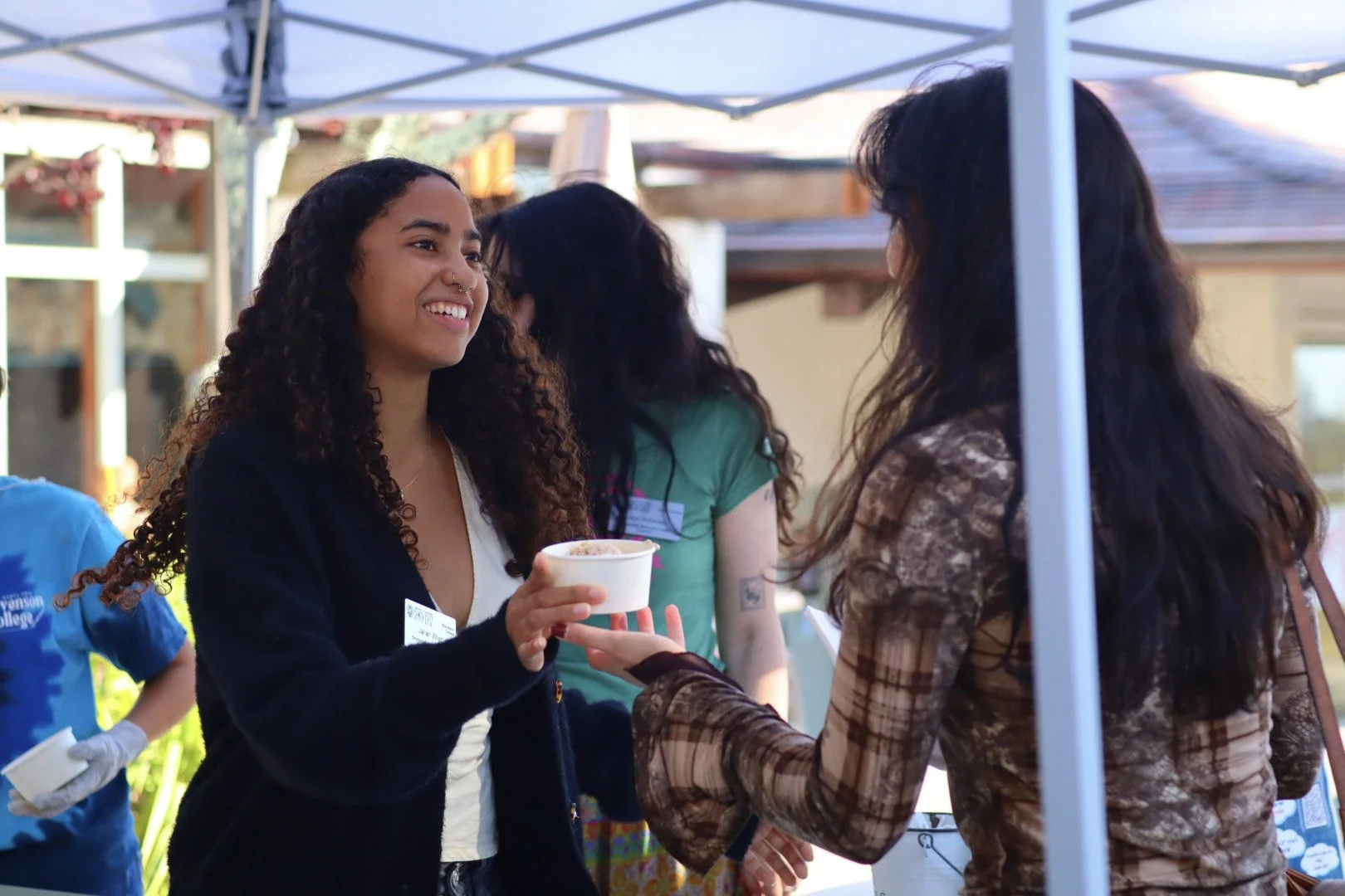 Two women are engaged in a conversation at an outdoor event, one holding a small bowl and the other reaching to take it. They are under a canopy with other people in the background.
