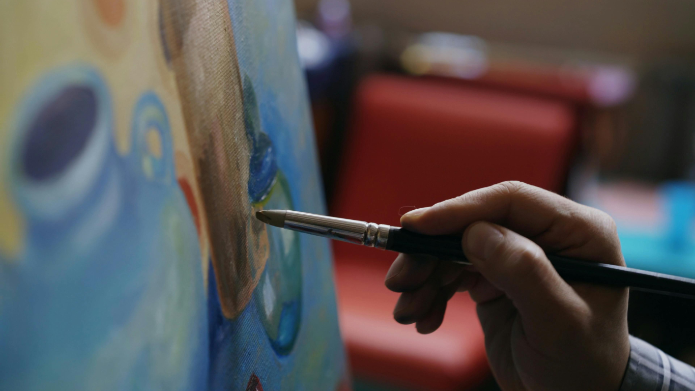 Close-up of an artist's hand holding a paintbrush, painting on a canvas with colorful abstract shapes.