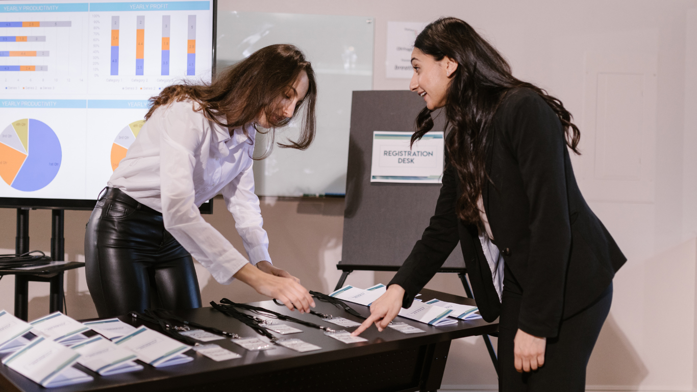 Two women at a registration desk, one pointing at conference badges or lanyards on a table, the other looking at them, in a professional setting with charts displayed on a screen behind them.