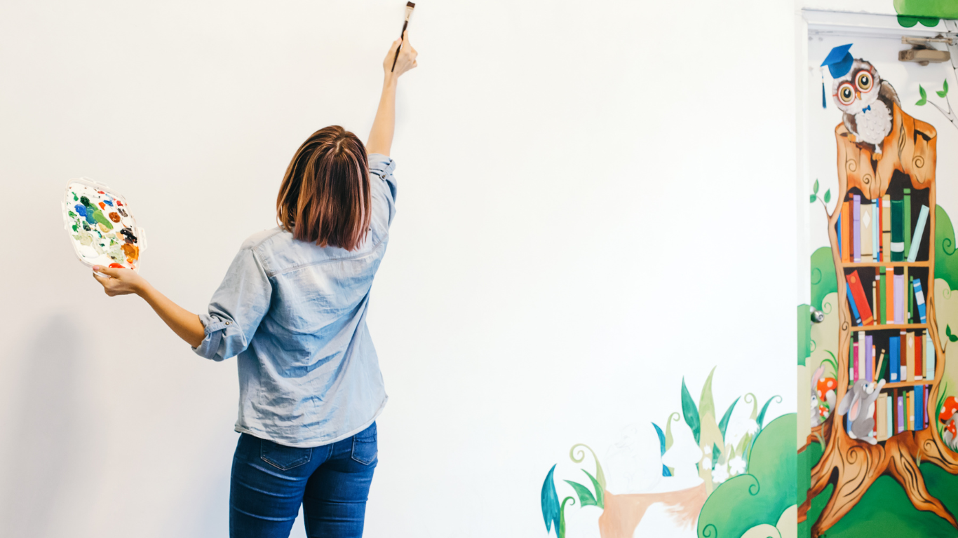 Person painting a mural of an owl on a wall next to a bookshelf in a colorful, whimsical style.