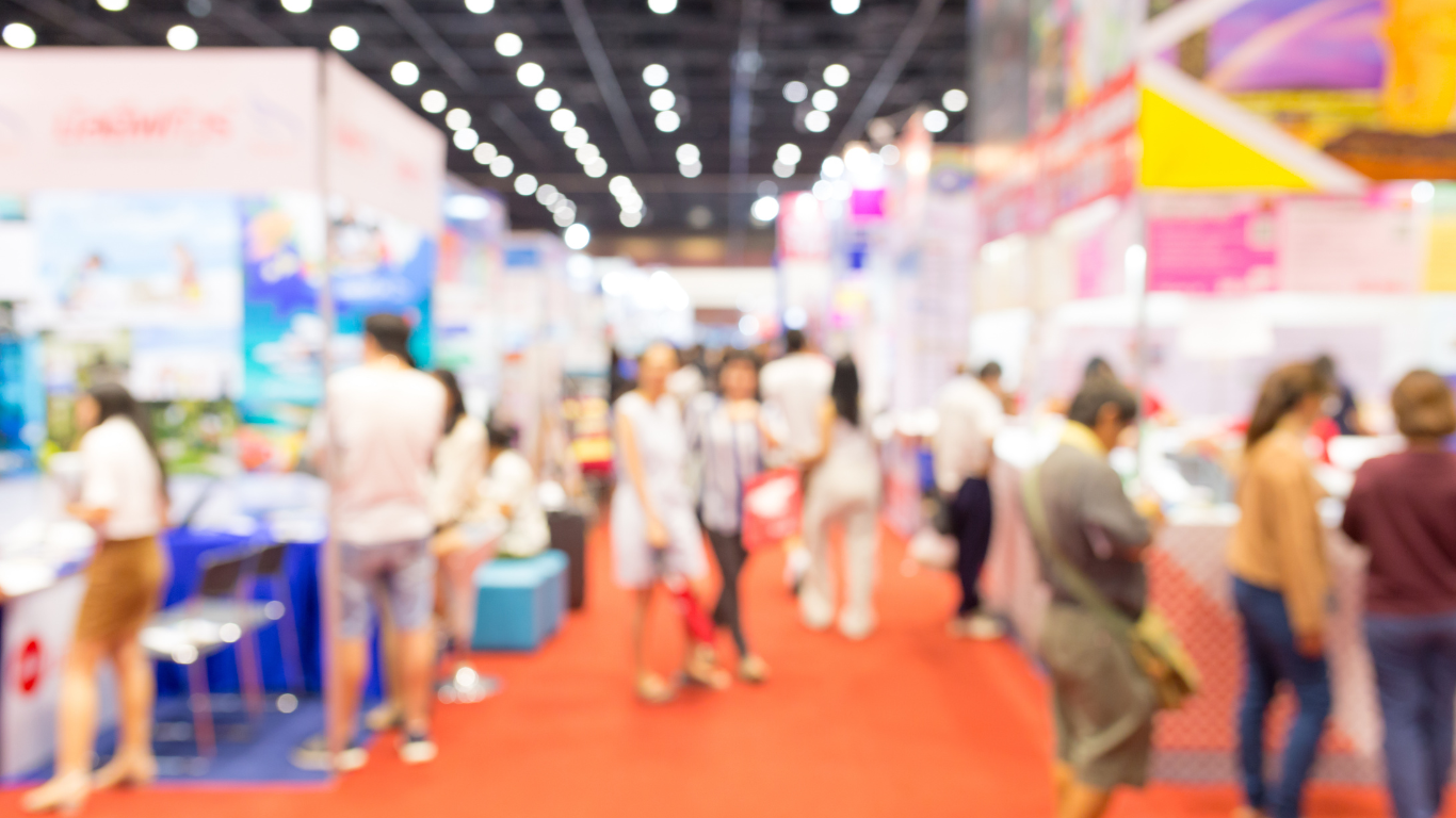 Blurred image of an indoor event or exhibition with people walking and visiting various booths and displays.