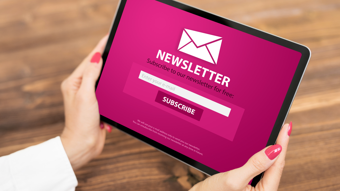 Tablet displaying a pink newsletter subscription form with a mail icon, a field to enter email, and a subscribe button, held by a person with pink nail polish over a wooden surface.
