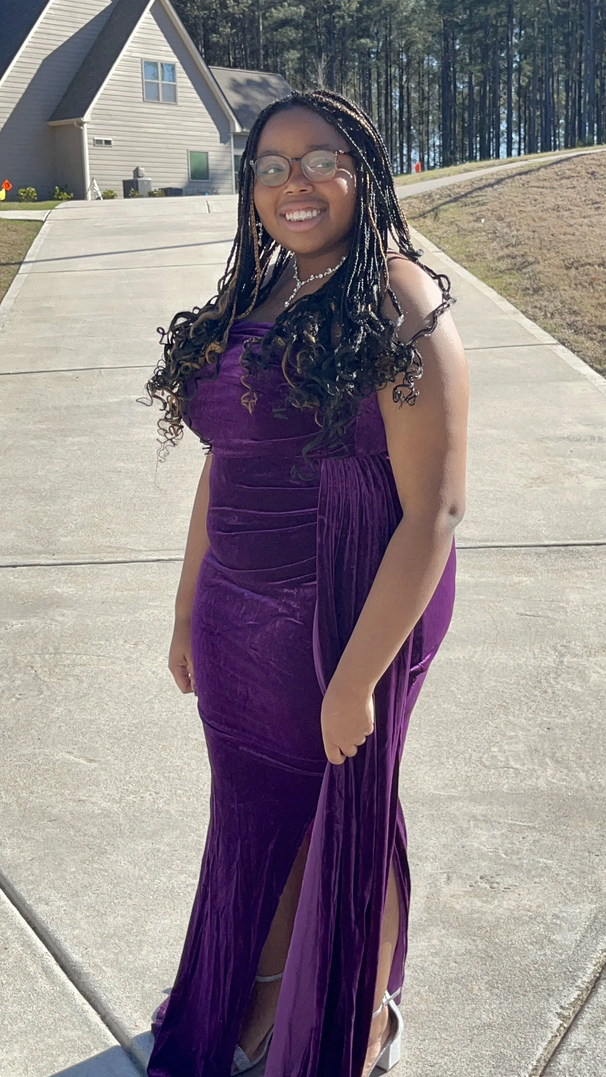 A young woman with glasses and long braided hair smiling outdoors on a sunny day, wearing a purple velvet dress with a slit, standing on a concrete sidewalk with a house and trees in the background.
