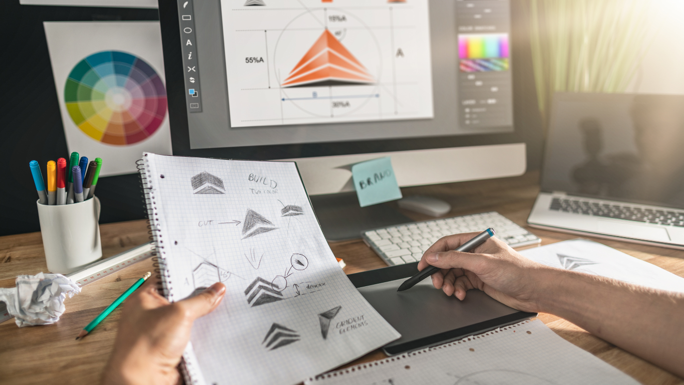 A person sketches geometric diagrams and notes on graph paper with a digital pen tablet at a creative workspace. There is a drawing of a building structure, arrows indicating construction steps, along with labeled notes such as 'BUILD' and 'CIRCULAR ARCHES.' In the background, a computer monitor displays a colorful graph, and a collection of colored pens is in a white cup on the wooden desk.