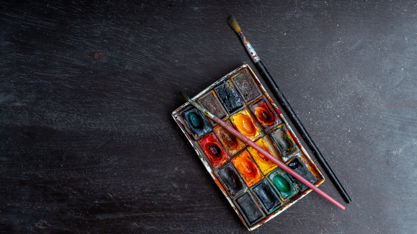 A watercolor paint palette with multiple colors, two paintbrushes, all resting on a dark wooden surface.