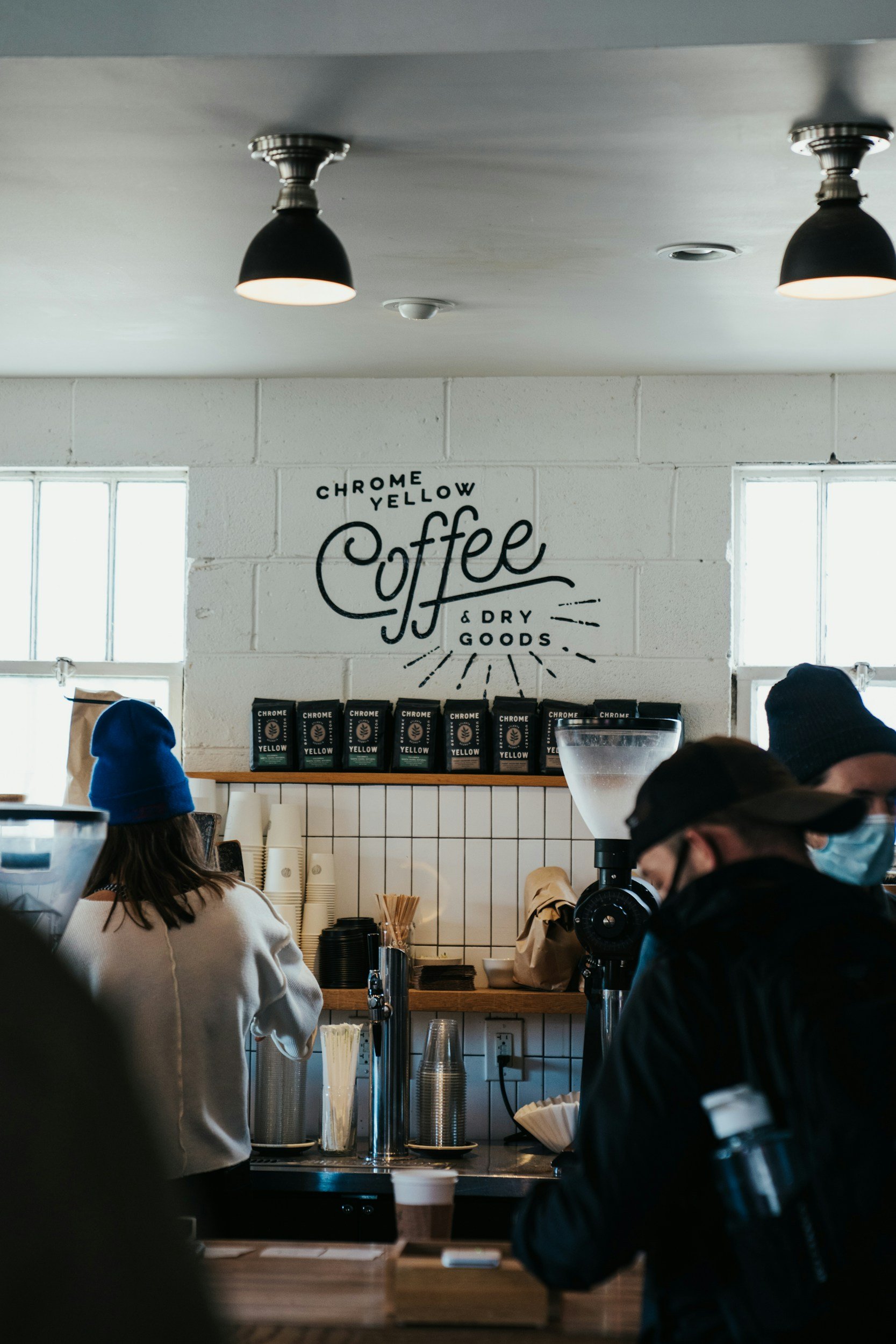 Inside a coffee shop, people queueing at the counter, with a sign on the wall reading 'Chrome Yellow Coffee & Dry Goods.'