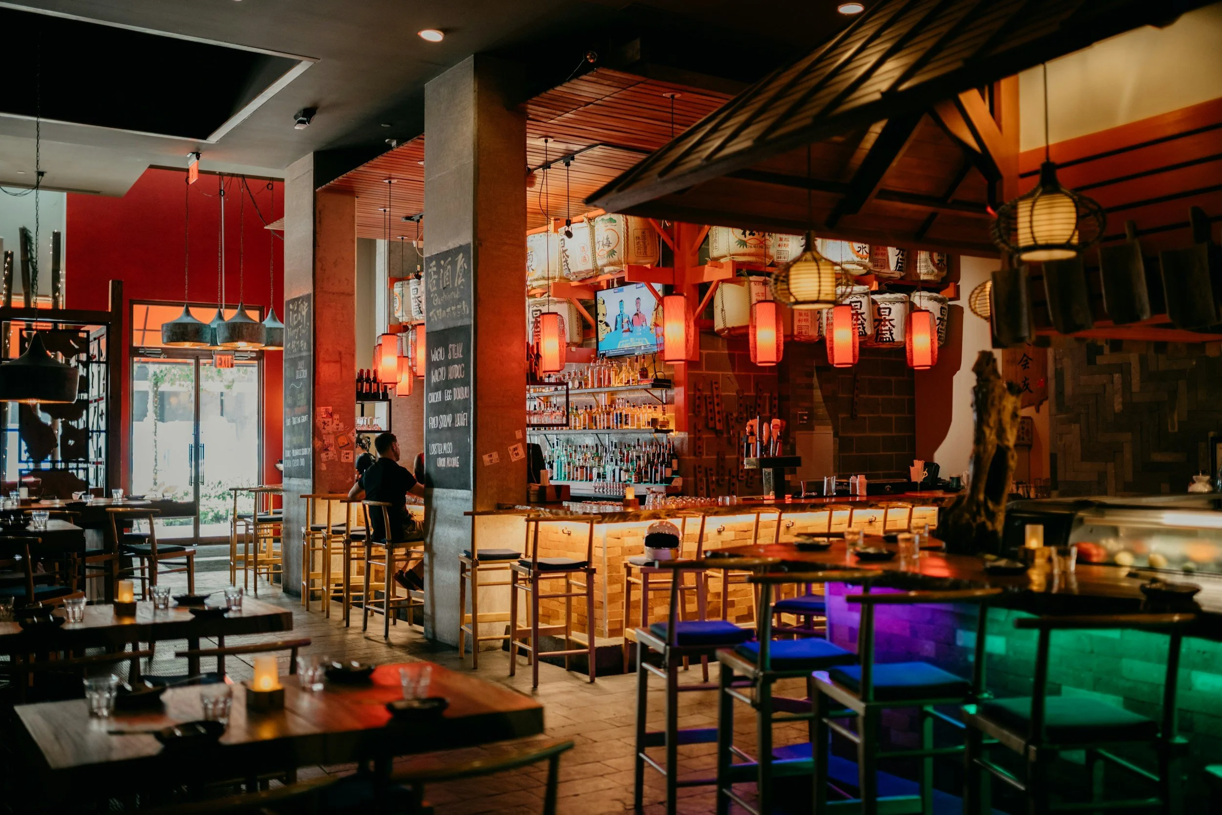 Interior of a restaurant or bar with wooden and brick decor, hanging lanterns, a TV screen, a bar area with bottles, and tables set for dining