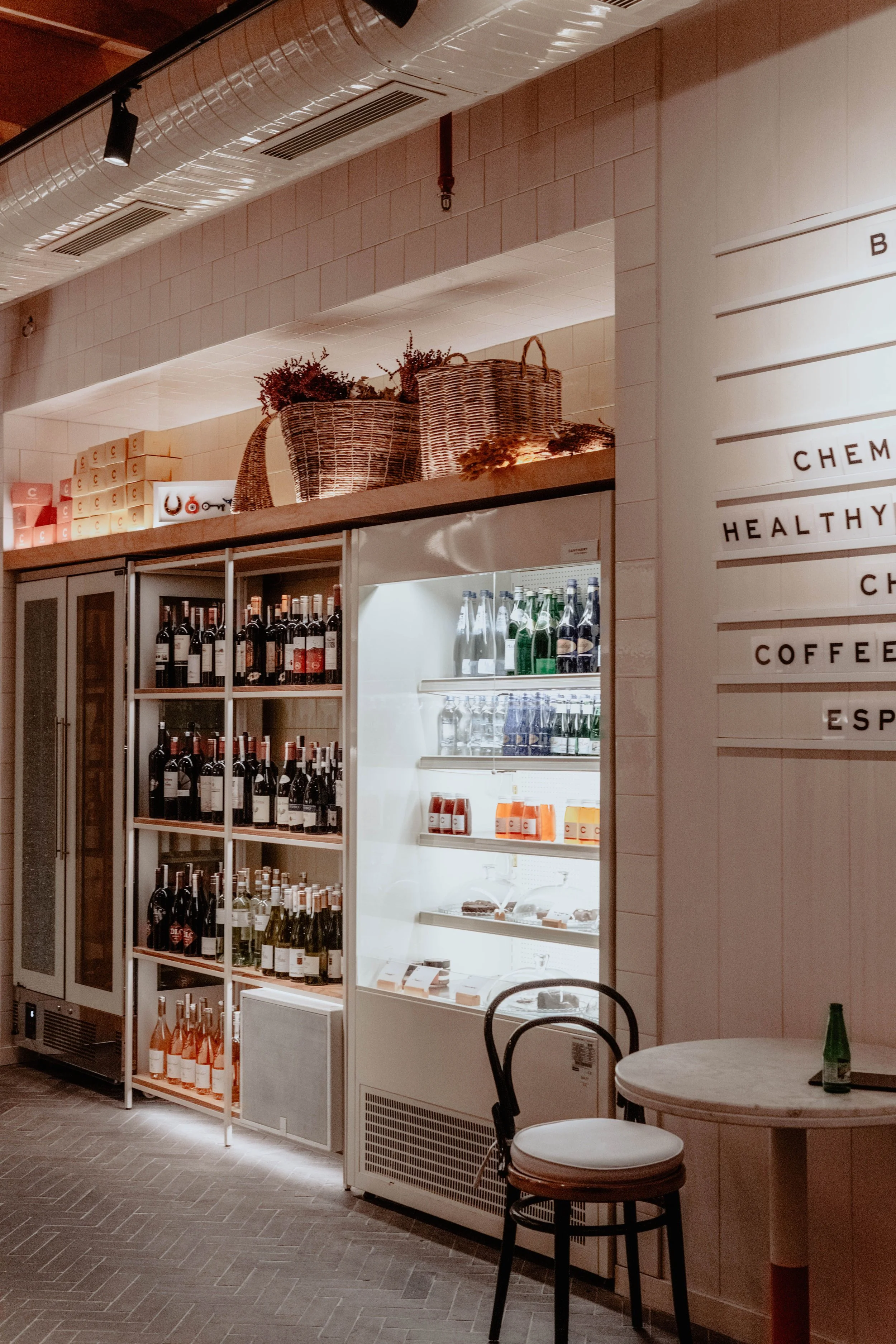 Refrigerator stocked with bottles of wine and beverages, with baskets on top, in a cafe or restaurant setting with a round table and chairs in the foreground.