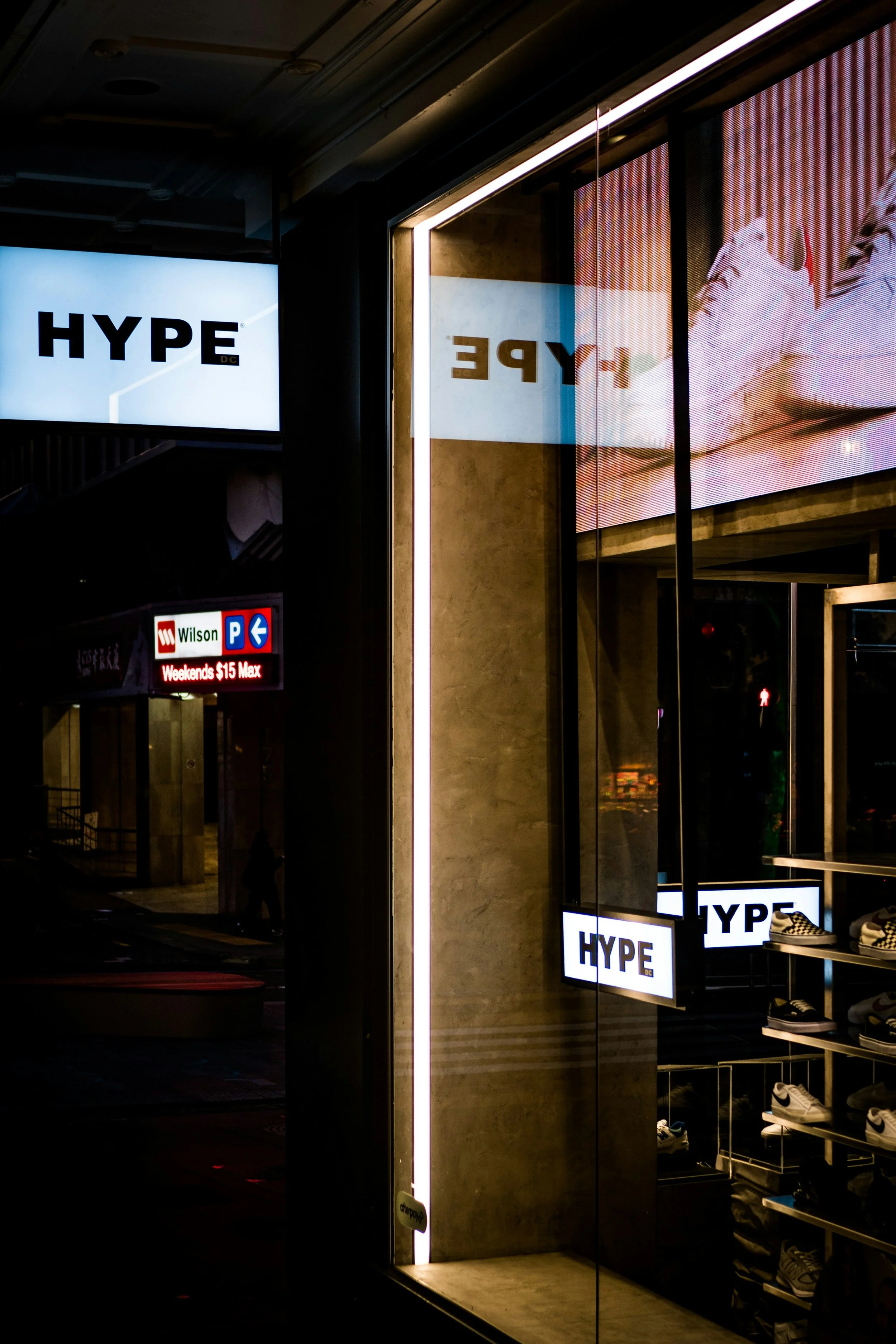 Night view of a store window display with illuminated signs reading 'HYPE' and showing shoes. The reflection in the window reveals part of a city street and a person walking by.