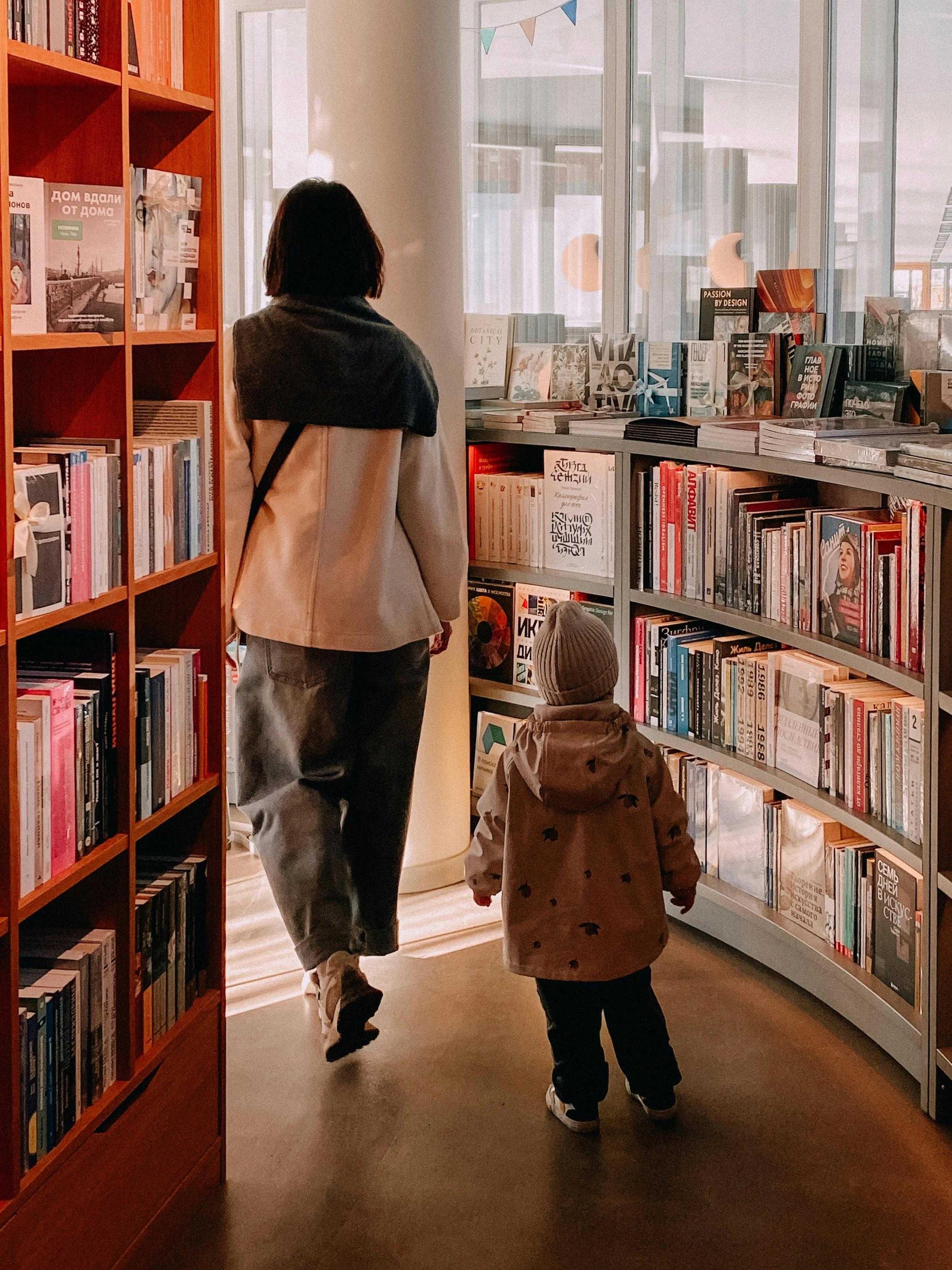 A woman and a young child walk through a bookstore. The woman is on the left, wearing a beige coat and jeans, with dark hair. The child is on the right, wearing a brown jacket with black stars and a gray beanie. The bookstore has bookshelves filled with various books and large windows letting in natural light.
