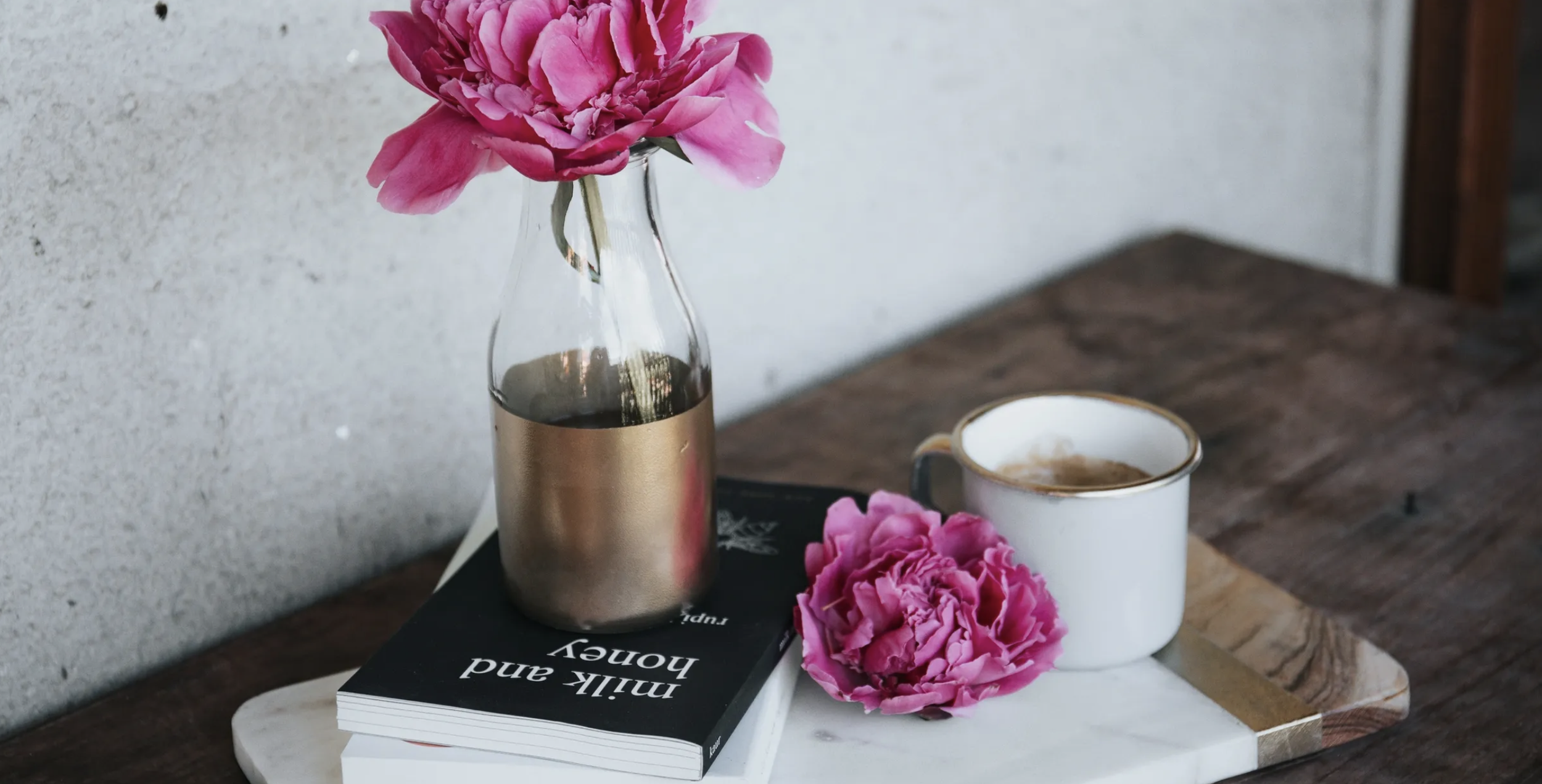 A pink peony flower in a clear glass vase with a gold band, placed on a black book titled 'think and grow rich'. Next to it, a pink peony flower on a white marble tray, with a white coffee mug with a gold rim, filled with coffee, on a wooden table.