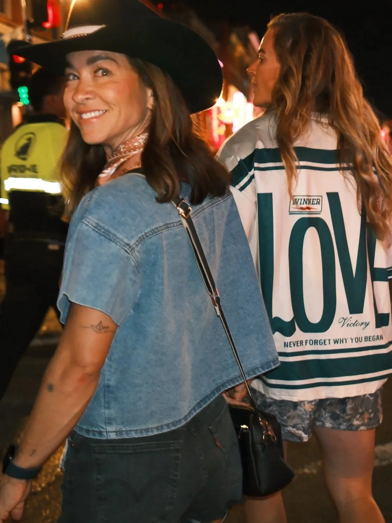 Two women standing at night, one smiling and wearing a cowboy hat, denim jacket, and black pants, the other with long red hair, wearing a large jersey with the words "LOVE," a jacket, and shorts, in a lively street scene with a police officer in the background.