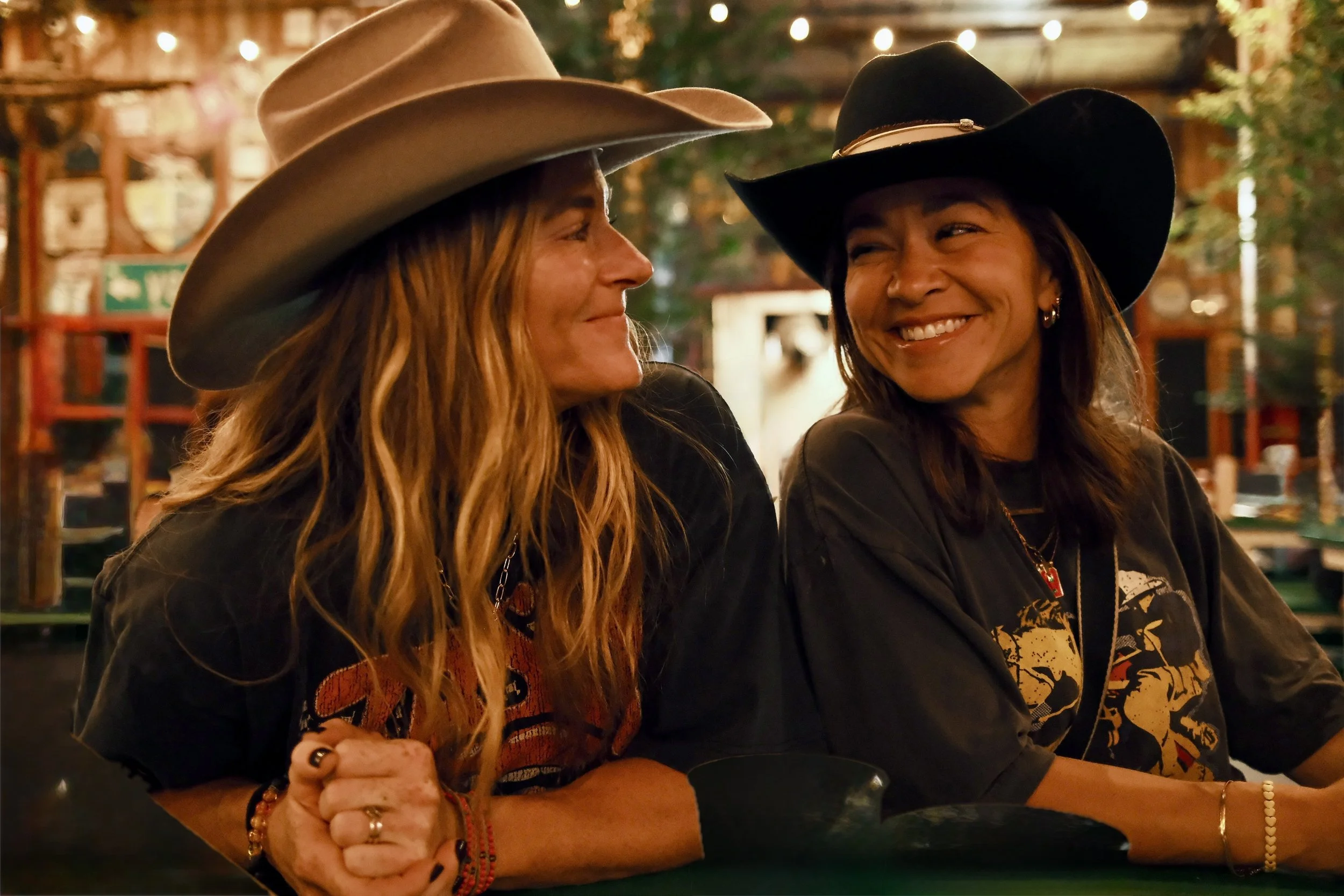 Two women wearing cowboy hats sitting together at a warmly lit indoor or outdoor setting, smiling and engaging in conversation.