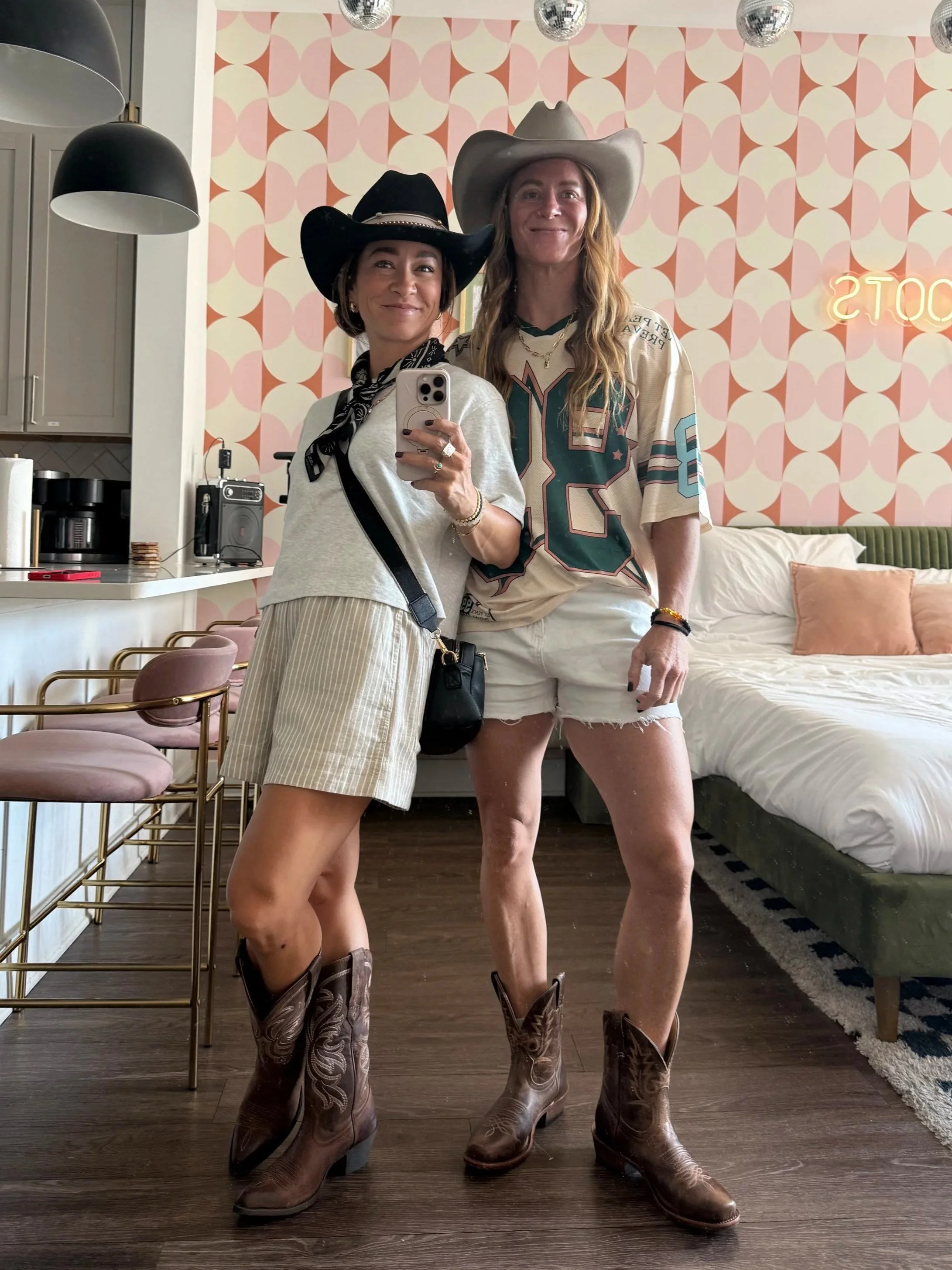 Two women dressed in country western style cowboy hats, boots, and casual clothing, standing in a room with pink patterned wallpaper, taking a selfie in front of a mirror.