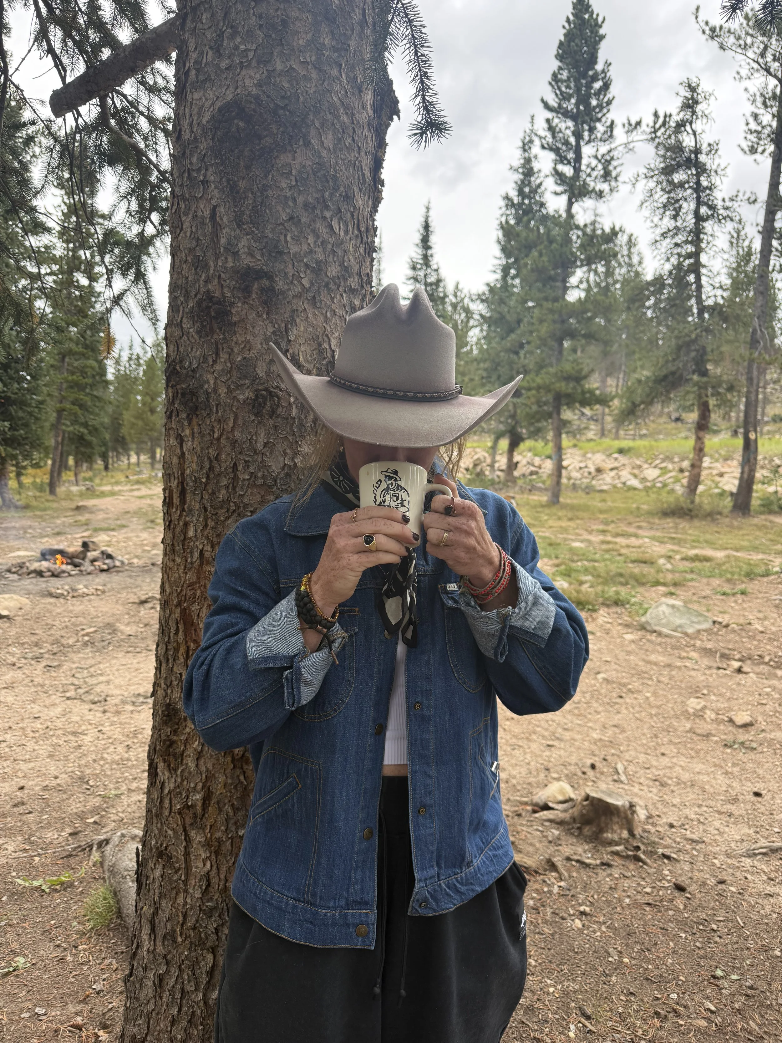 Person standing outdoors next to a tree, wearing a wide-brimmed hat, denim jacket, and holding a coffee mug in front of their face, with a forested background and a campfire visible in the distance.