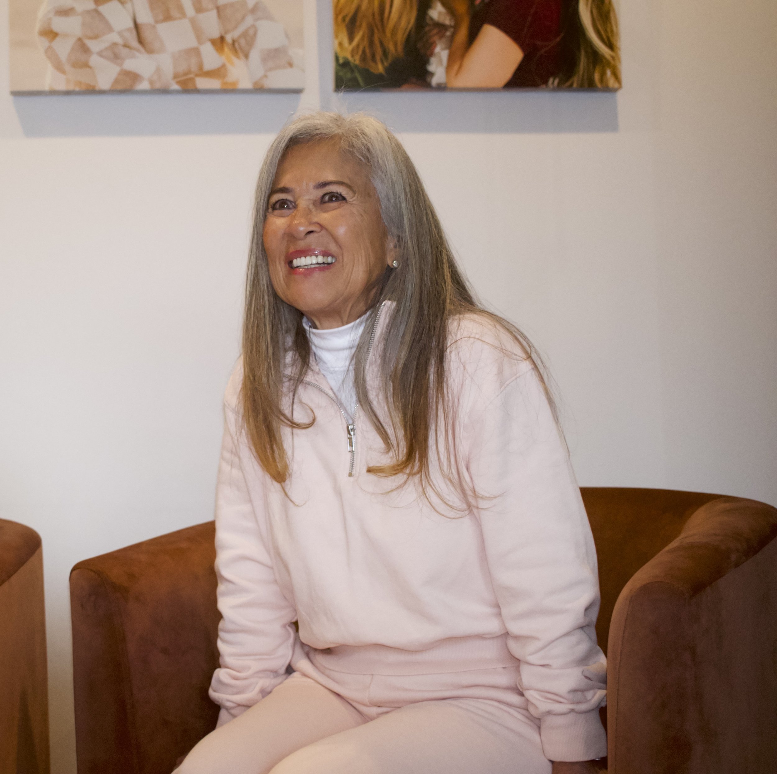 A woman with long gray hair smiling, seated in a corner with a wooden wall behind her. She is wearing a light pink jacket and cream pants. There are framed photos above her, including one of a girl with long hair.