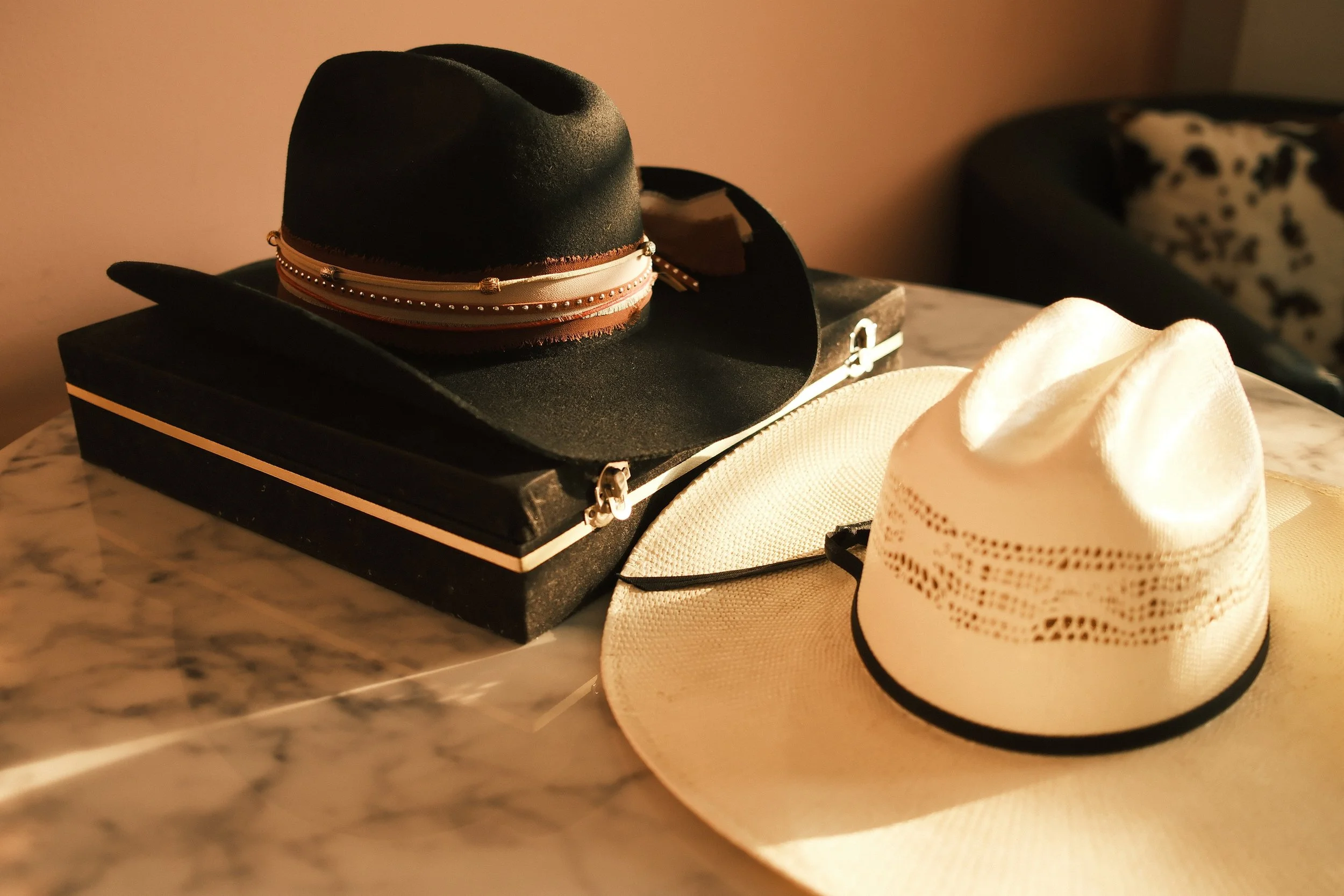 A black hat with a wide brim, adorned with beaded bands, resting on a black case on a marble surface. Next to the hat is a white hat with a wide brim and black decoration, and an out-of-focus patterned object or fabric in the background.