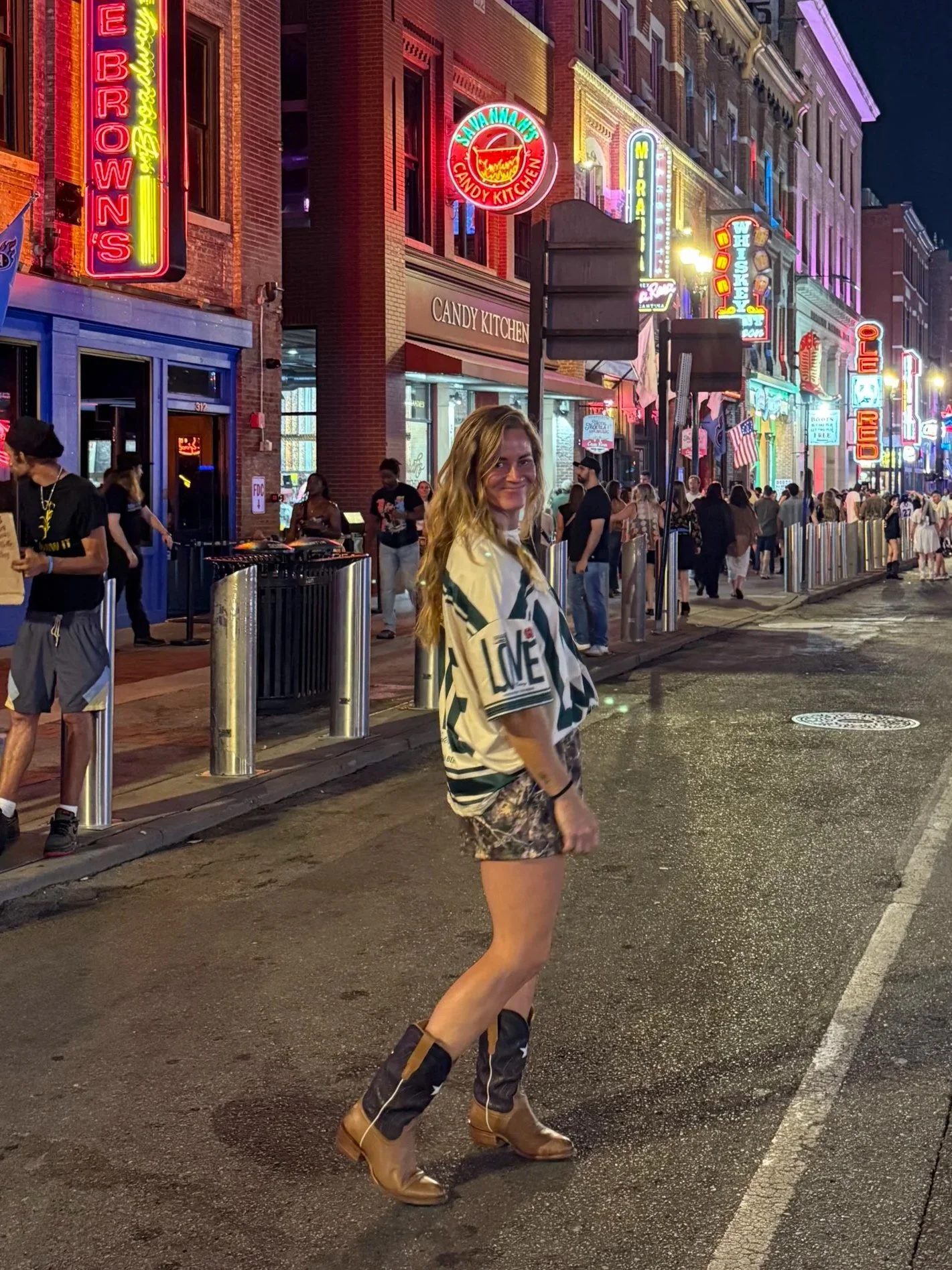 A young woman with long, curly blonde hair wearing a loose, patterned shirt, camouflage shorts, and tan cowboy boots standing on a city street at night. Behind her are neon signs for various restaurants and bars, and a crowd of people walking along the sidewalk.