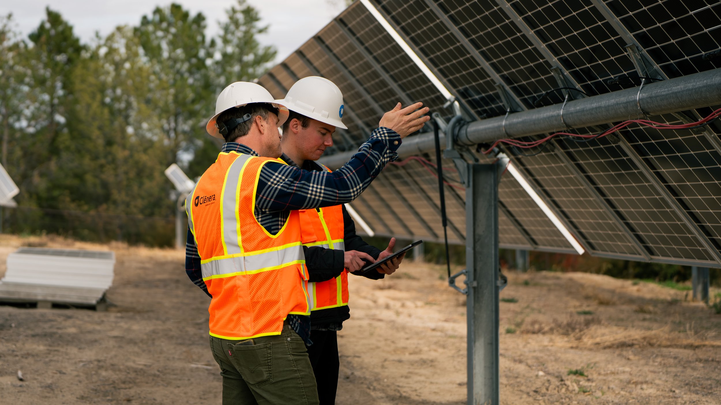 Two workers wearing hard hats and safety vests inspecting solar panels at a solar farm.
