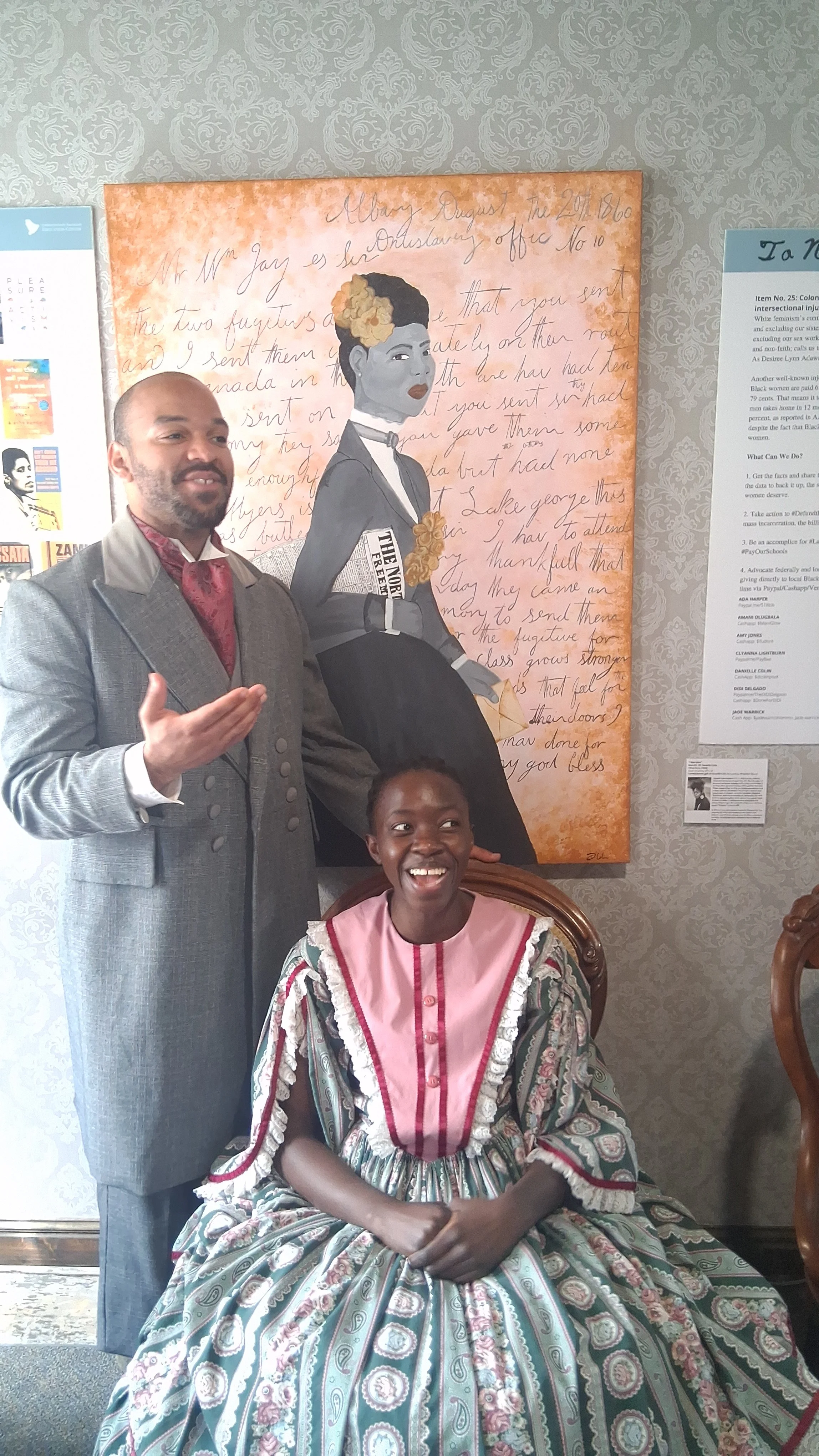 A man and a young girl sitting and standing in front of a large mural of an illustrated woman with flowers in her hair, with handwritten text in the background. The man is wearing a gray suit and the girl is wearing a patterned dress with pink accents, both smiling.