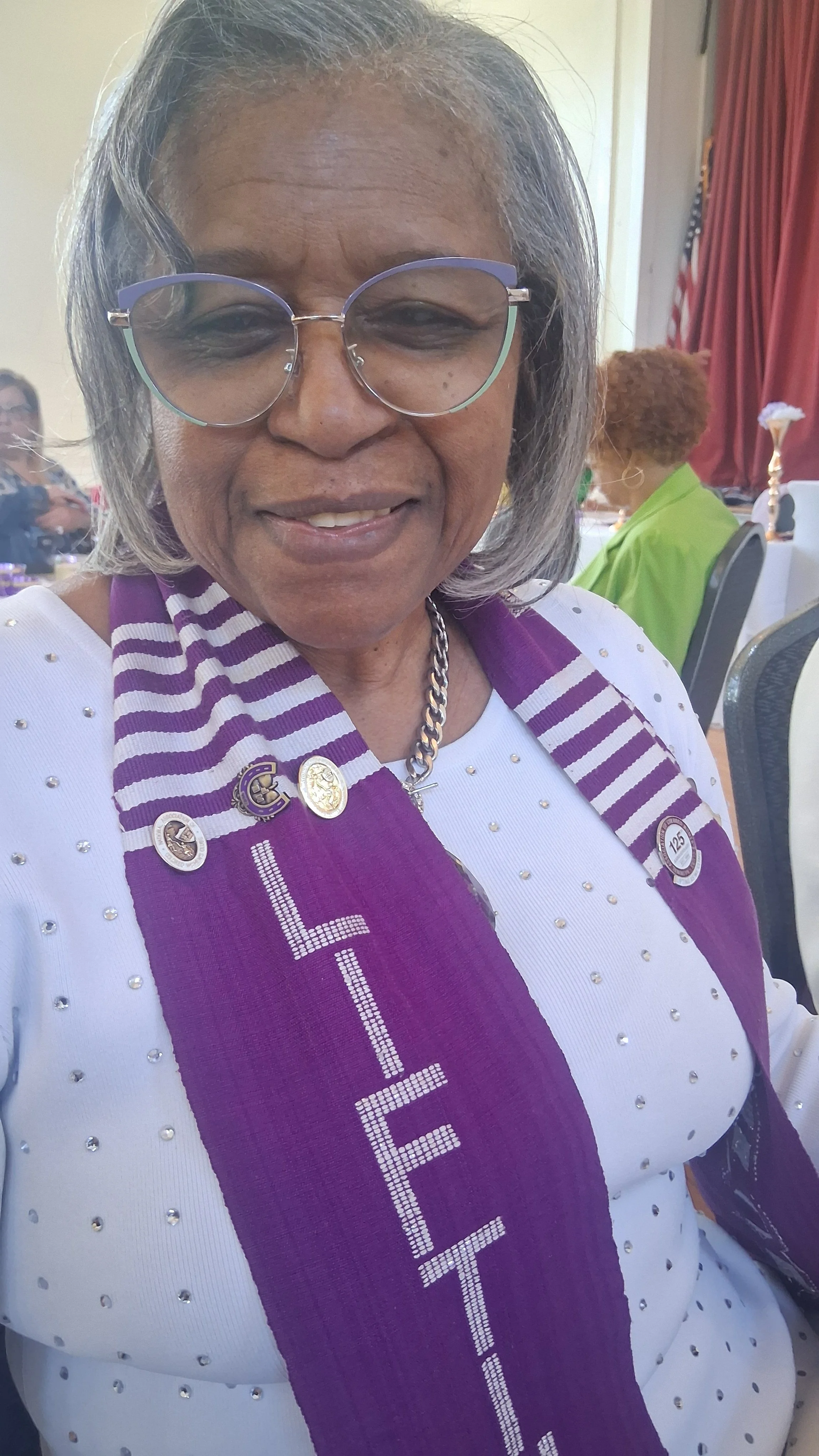 An older woman with gray hair and glasses smiling at a celebration or event. She is wearing a white dress with small embellishments and a purple sash with the word 'ELETE' on it, along with pins and a chain necklace. In the background, other women and event decorations are visible.