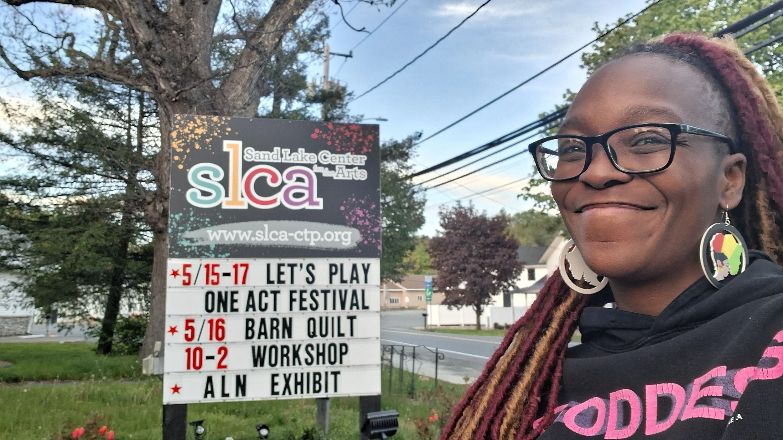 A woman with glasses and multicolored dreadlocks smiling in front of a sign for the Sand Lake Center for the Arts, with trees and houses in the background.