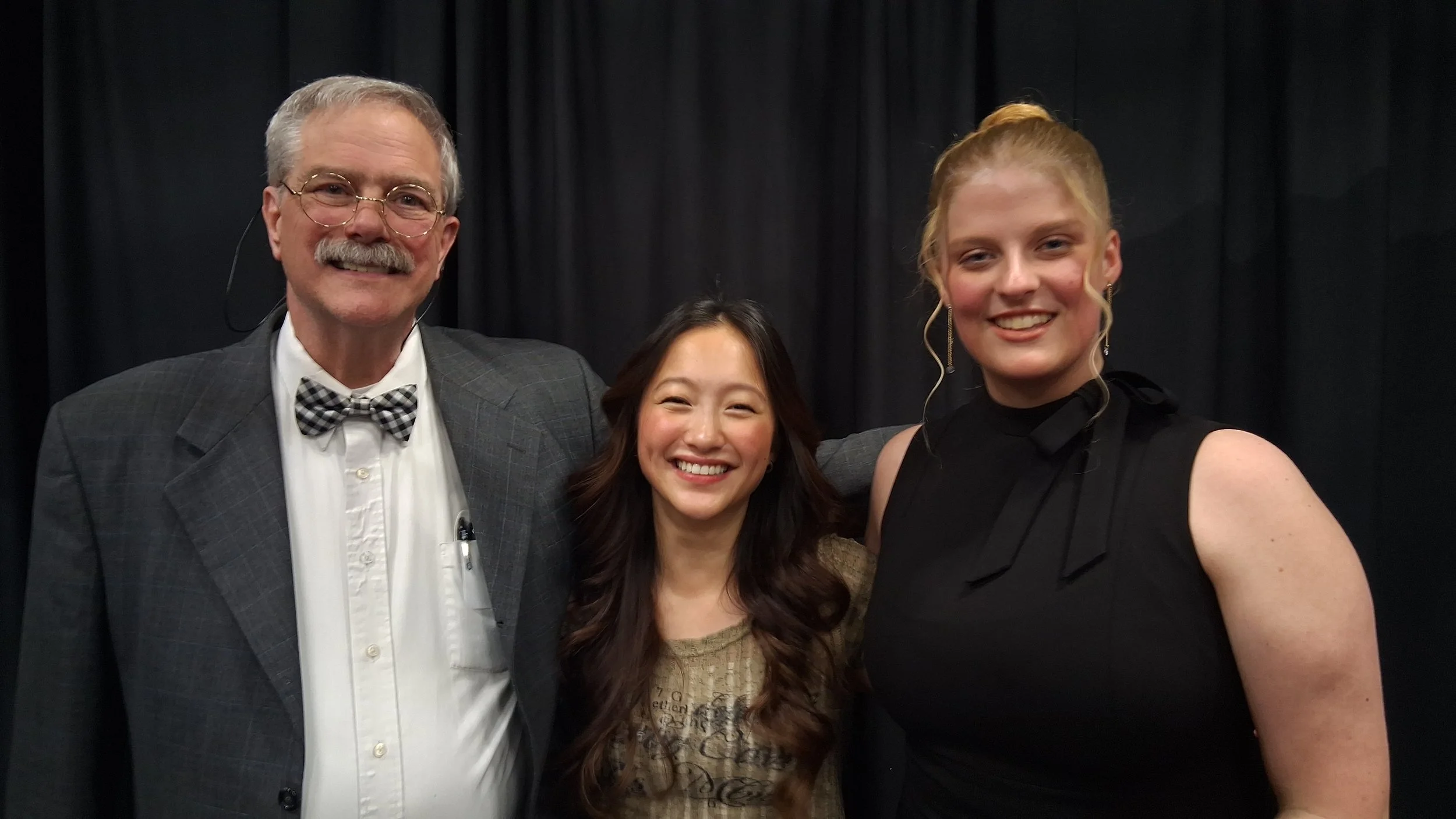 Two smiling women and one older man posing together in front of a black curtain background.