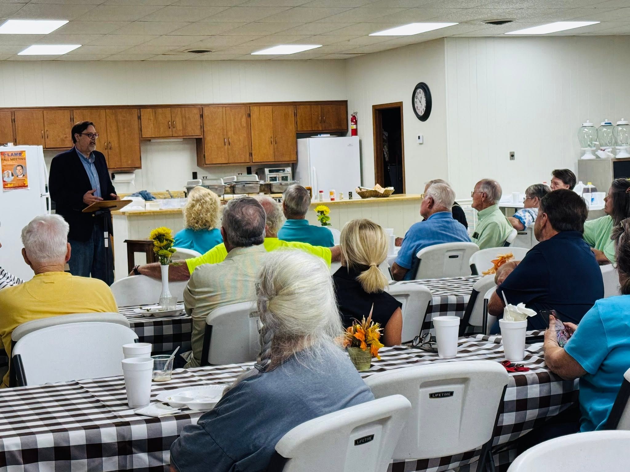 A speaker giving a presentation to an audience in a community hall kitchen with wooden cabinets and a clock on the wall.