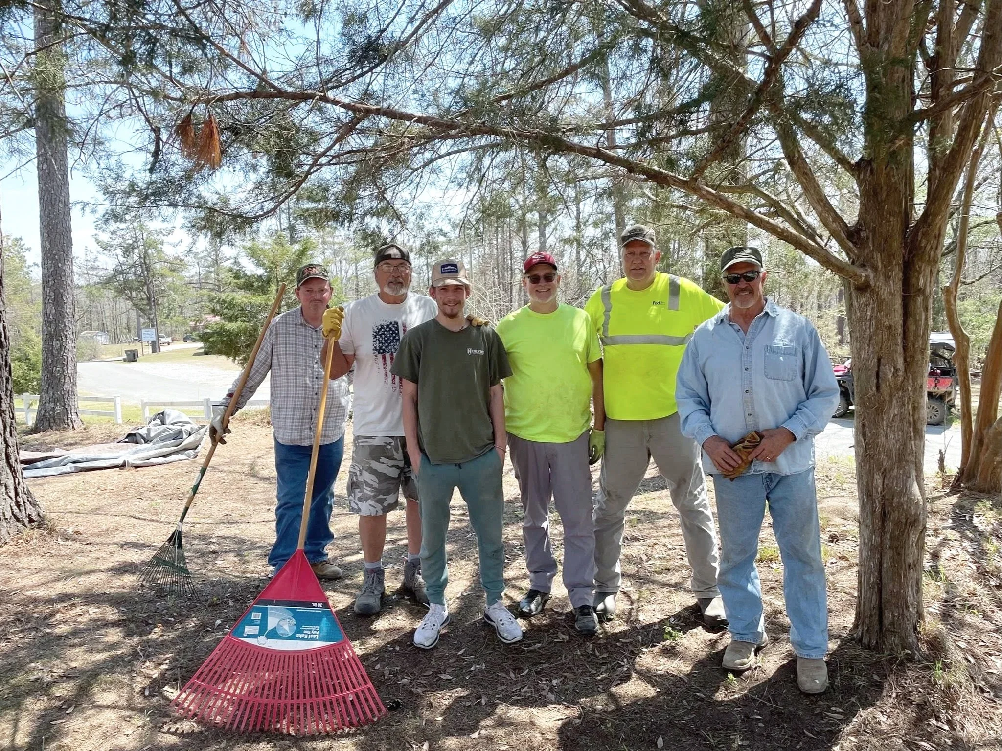 A group of seven men standing outdoors under a large tree during daytime. They are smiling and appear to be involved in community or environmental work. Some men are holding gardening tools, and there are tools and supplies on the ground in front of them.