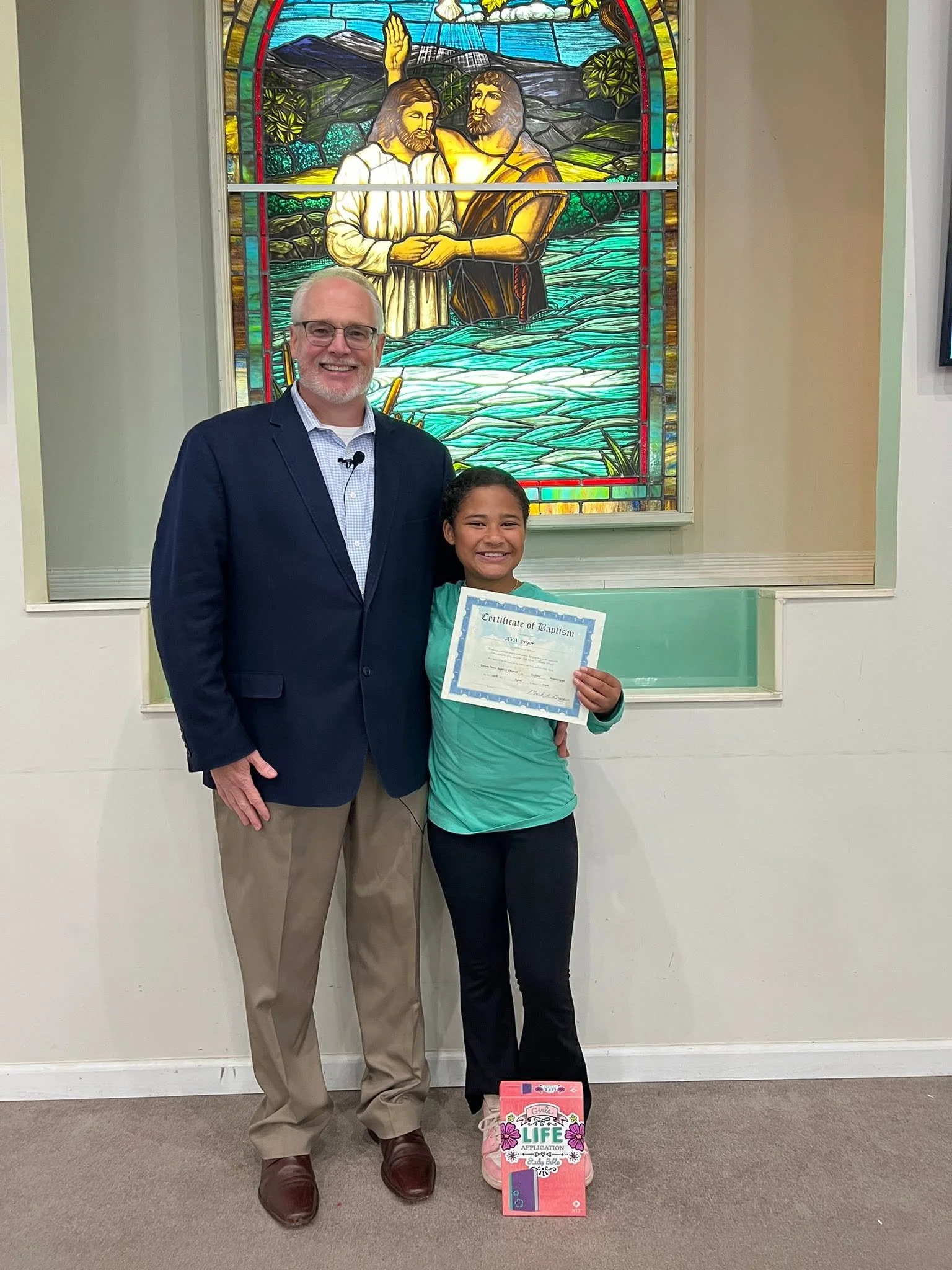 A man and a girl standing in front of a stained glass window depicting Jesus baptizing a man in a river. The girl is holding a certificate of baptism, and a pink gift bag with a 