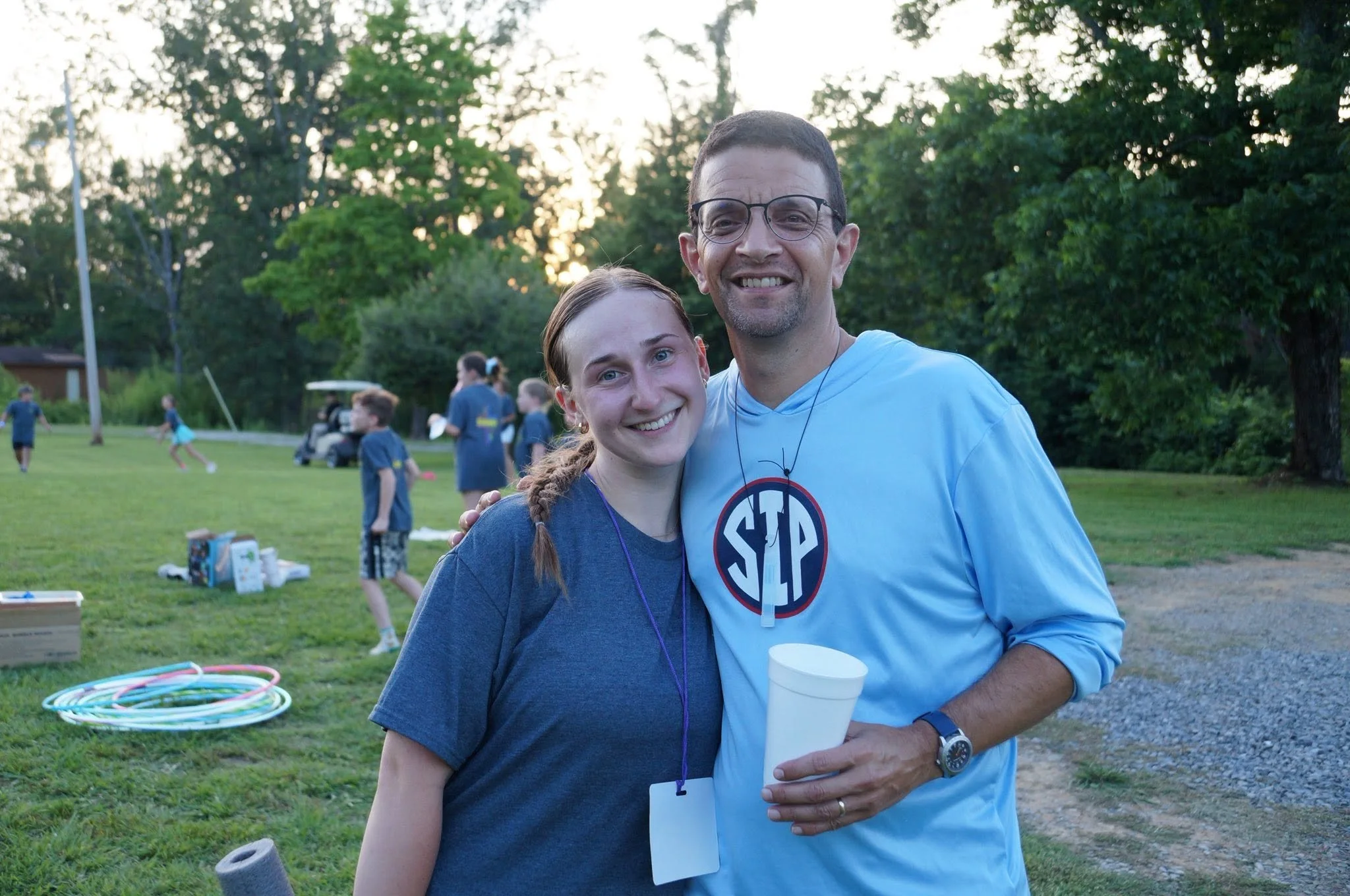 A smiling young woman and a man wearing glasses and a gray T-shirt with a logo are at a park during sunset. They are standing close together, with the woman holding a white cup. In the background, kids are playing, and various outdoor toys are scattered on the grass.