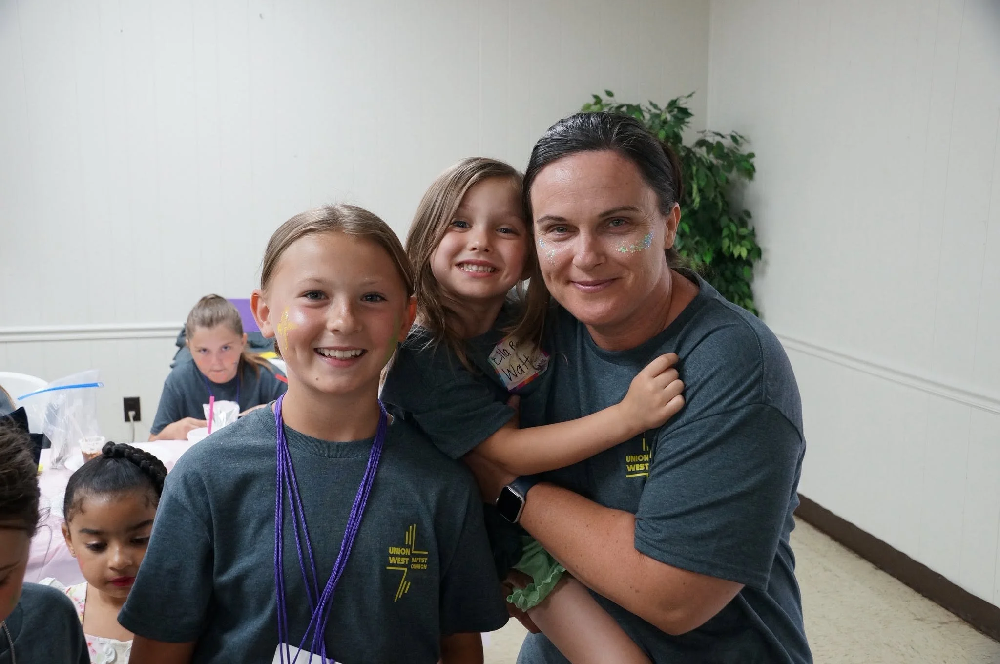 A group of children and an adult woman pose for a photo at an indoor event. The children have face paint and are wearing gray shirts with a yellow logo that says 'Union West Baptist Church.' The woman has face paint, is smiling, and hugging a girl. Other children are visible in the background, some sitting at a table with party favors and decorations.