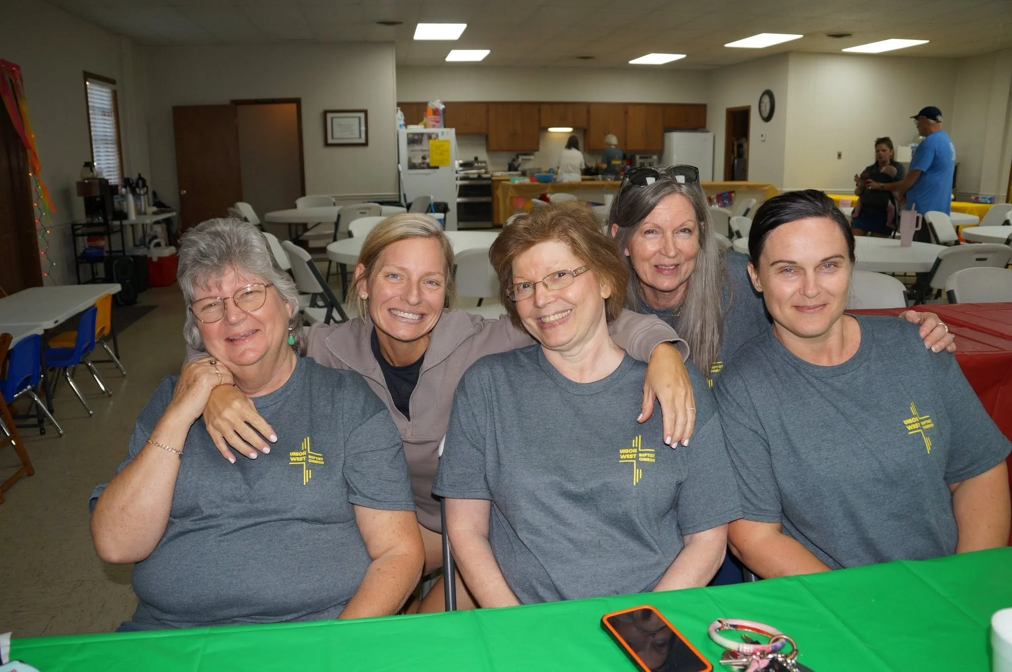 Group of five smiling women sitting at a table, wearing matching gray T-shirts with a yellow cross logo and text, in a community hall with tables, chairs, and kitchen area in the background.