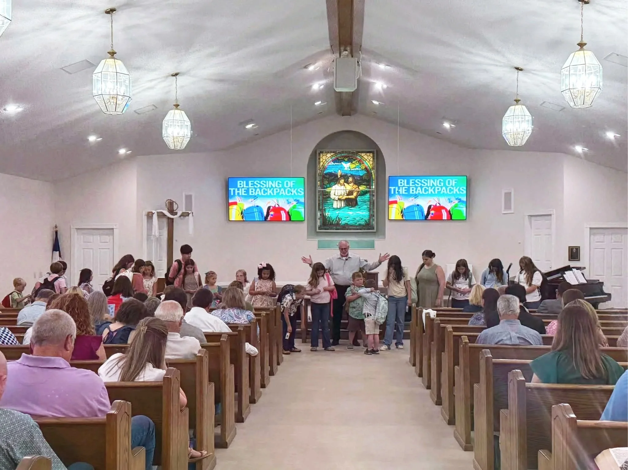 Inside a church with many people seated in wooden pews, a group of children and adults are gathered near the front, with a man standing and gesturing. There are two digital screens displaying 'Blessing of the Backpacks' on either side of a stained glass window depicting two figures and a dove.