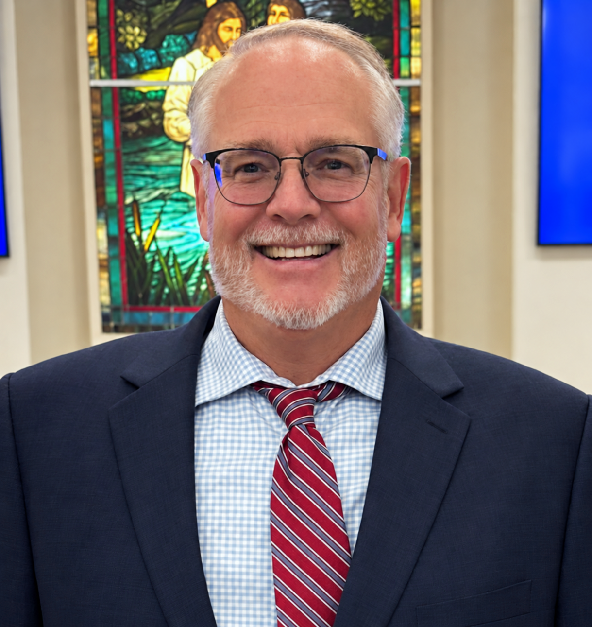 A smiling middle-aged man with glasses, wearing a dark suit, blue checkered shirt, and a red striped tie, standing in front of a stained glass window depicting Jesus.