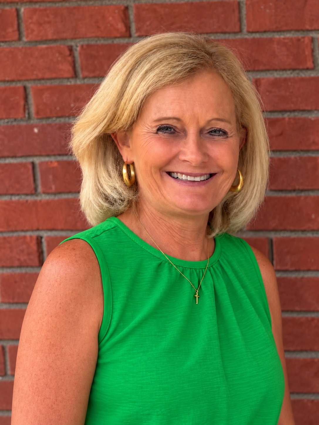 A smiling woman with blonde hair wearing a green sleeveless top, gold earrings, and a gold cross necklace, standing in front of a brick wall.