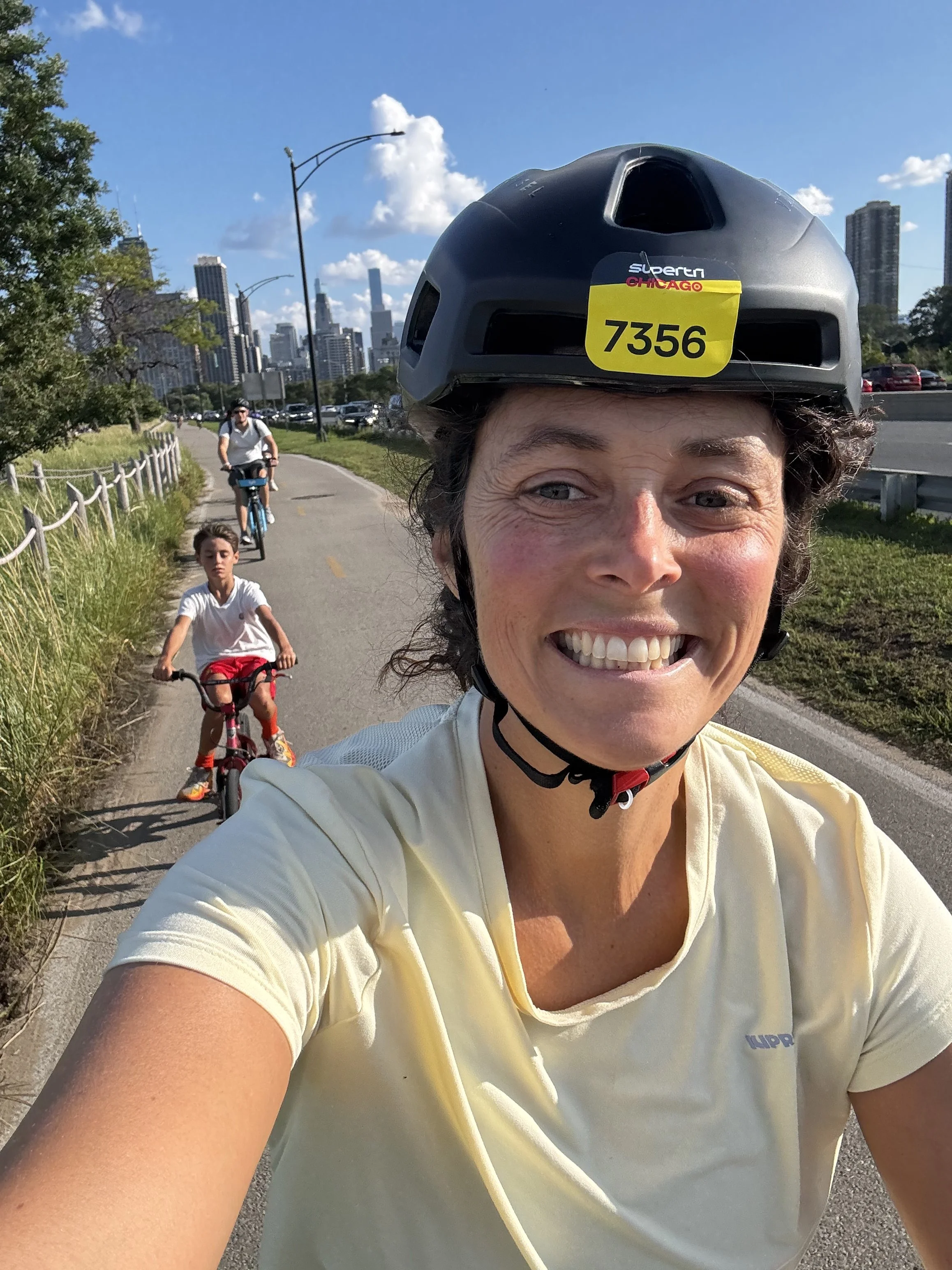 Une femme souriante portant un casque de vélo, prenant un selfie avec deux enfants en arrière-plan, à vélo sur une piste en bordure de ville.