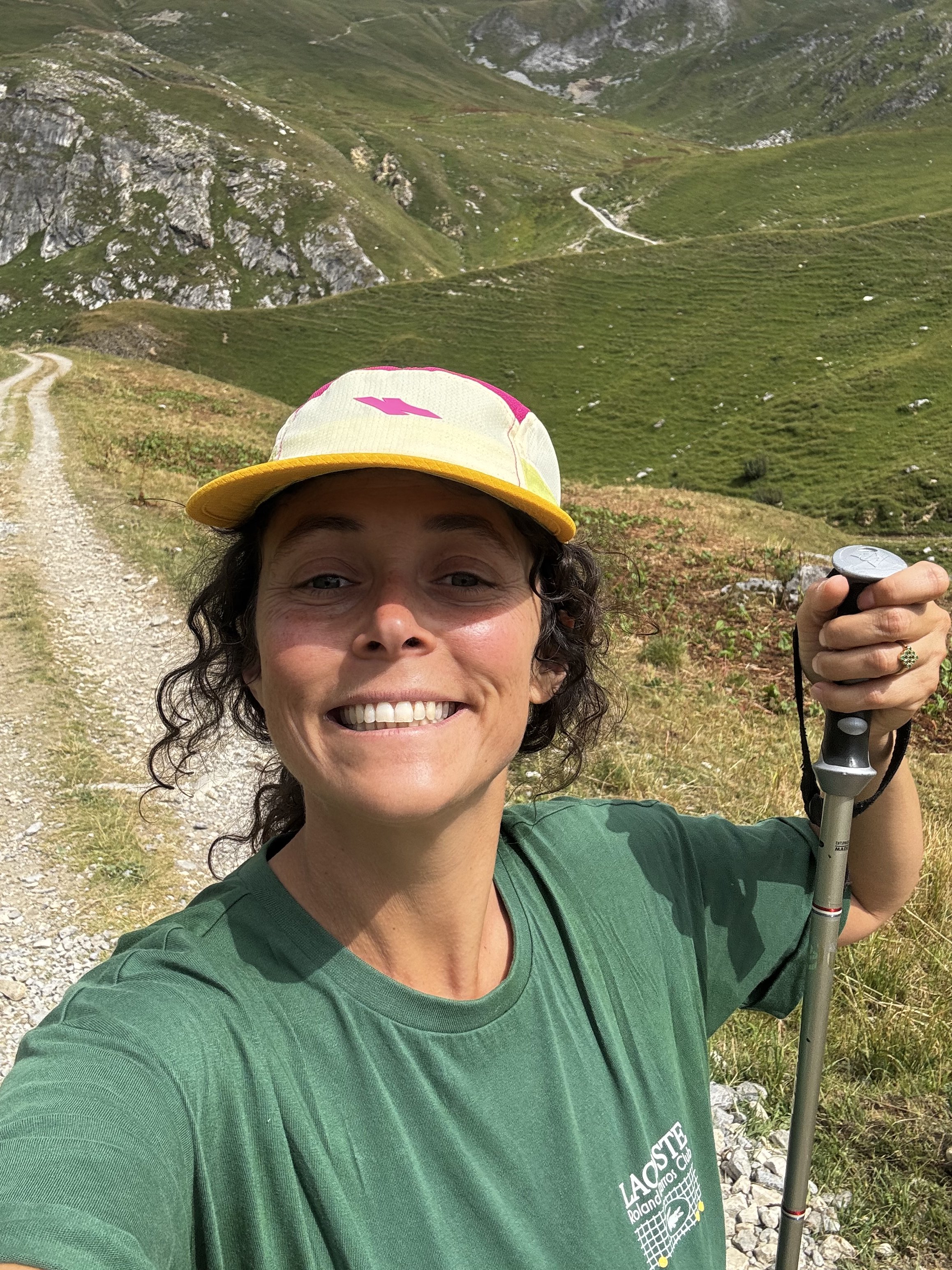 Jeune femme souriante portant un chapeau coloré, une veste verte, tenant une canne de marche, dans un paysage de montagnes verdoyantes avec un sentier en gravier.