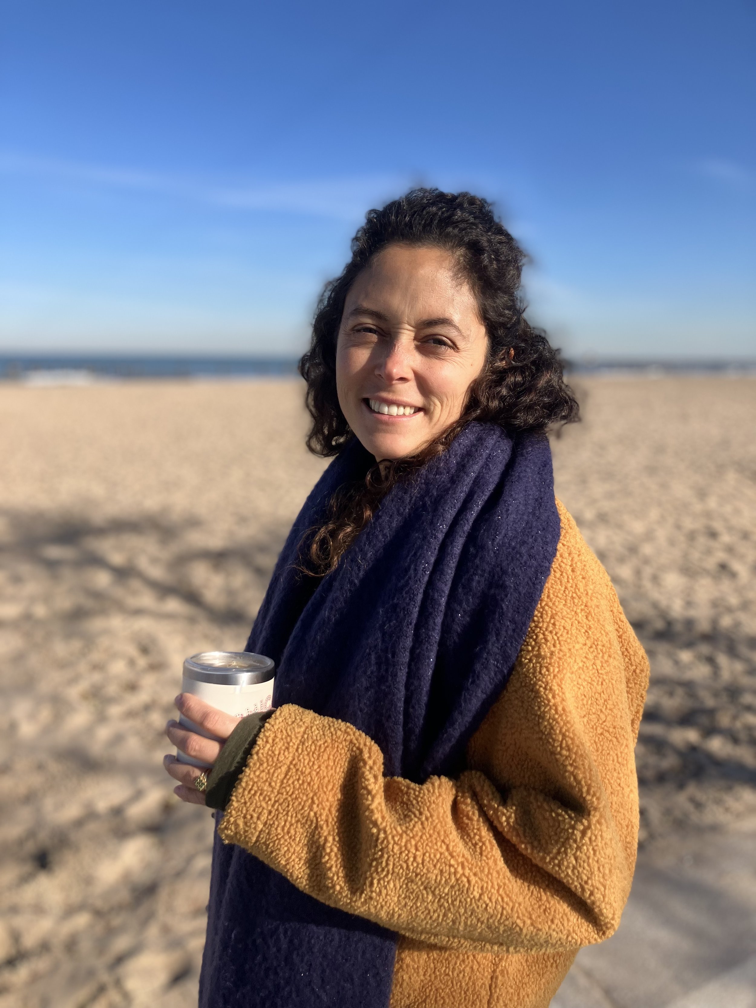 Femme souriante portant un manteau coloré avec un fond de plage et un ciel bleu.