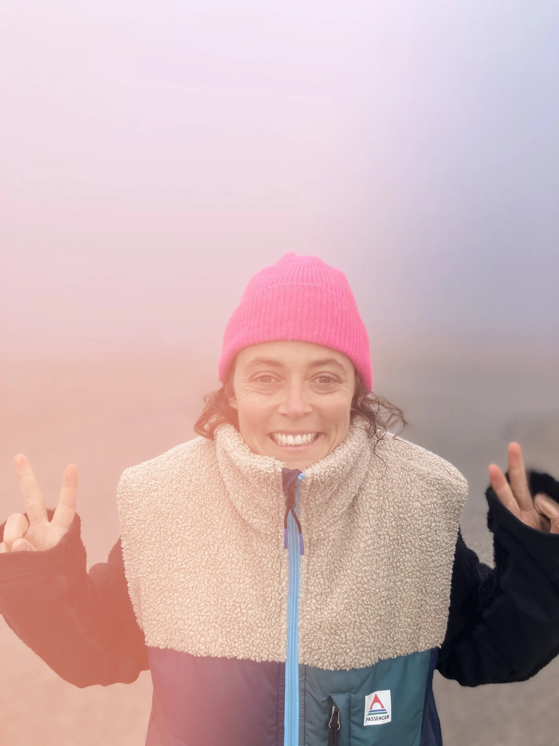 Une femme souriante portant un bonnet rose, un pull beige en peluche, et un manteau gris avec une étiquette PASSENGER, faisant un signe de paix avec deux doigts.