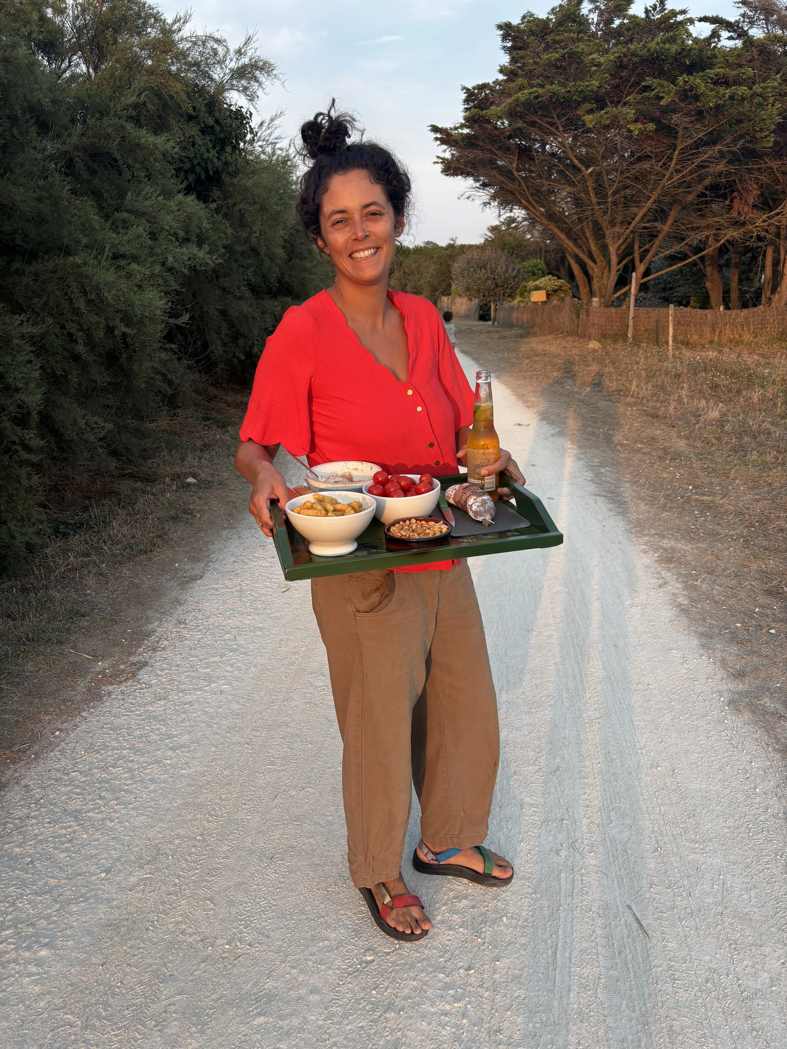 Femme souriante portant un haut rouge et un pantalon beige, tenant un plateau avec des plats et une boisson, devant un chemin de terre entouré d'arbres.