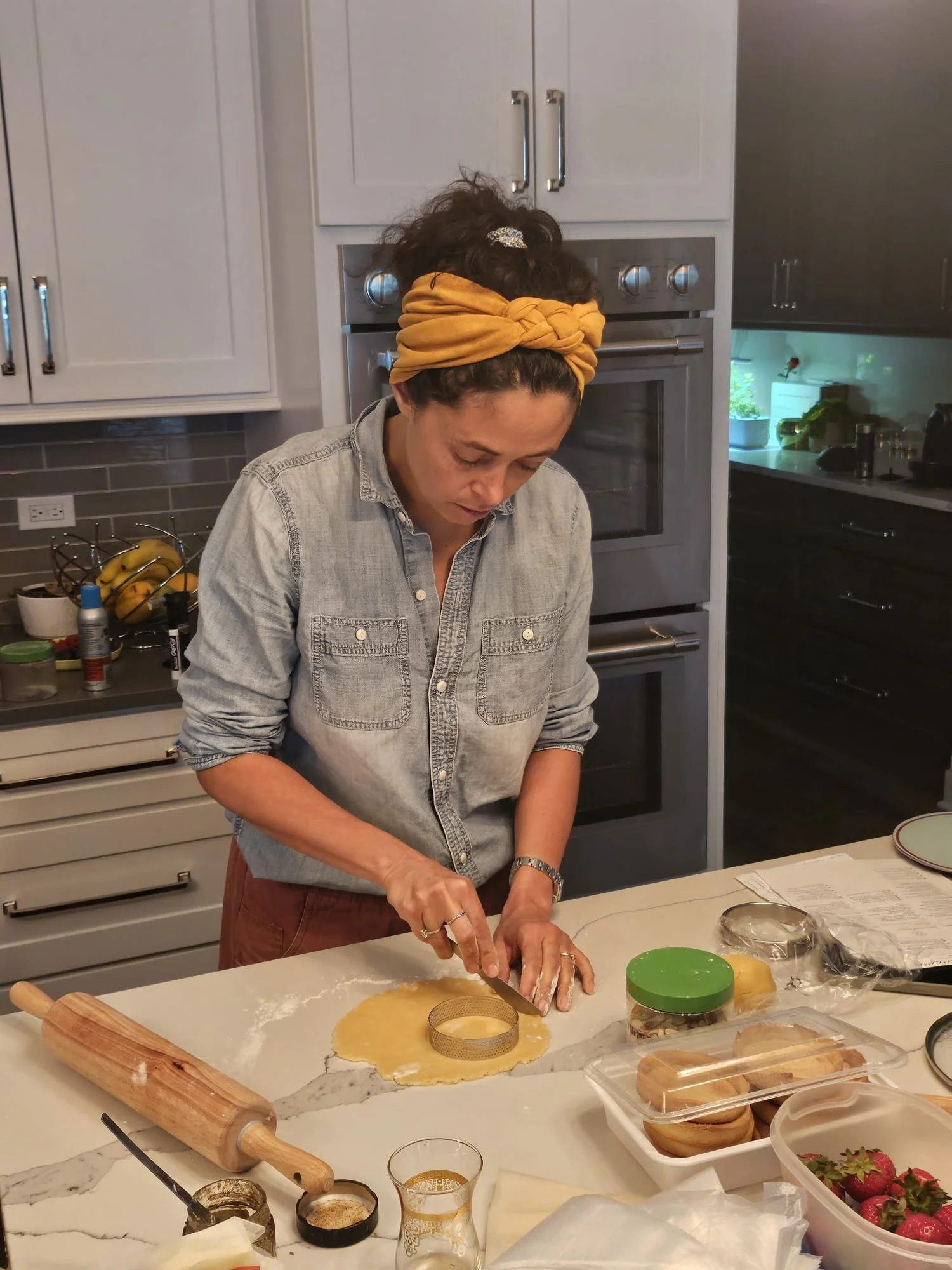 Une femme prépare des biscuits ou une pâte à l'aide d'un emporte-pièce rond, dans une cuisine moderne. Elle porte un bandeau jaune sur la tête, un jean gris et une montre.