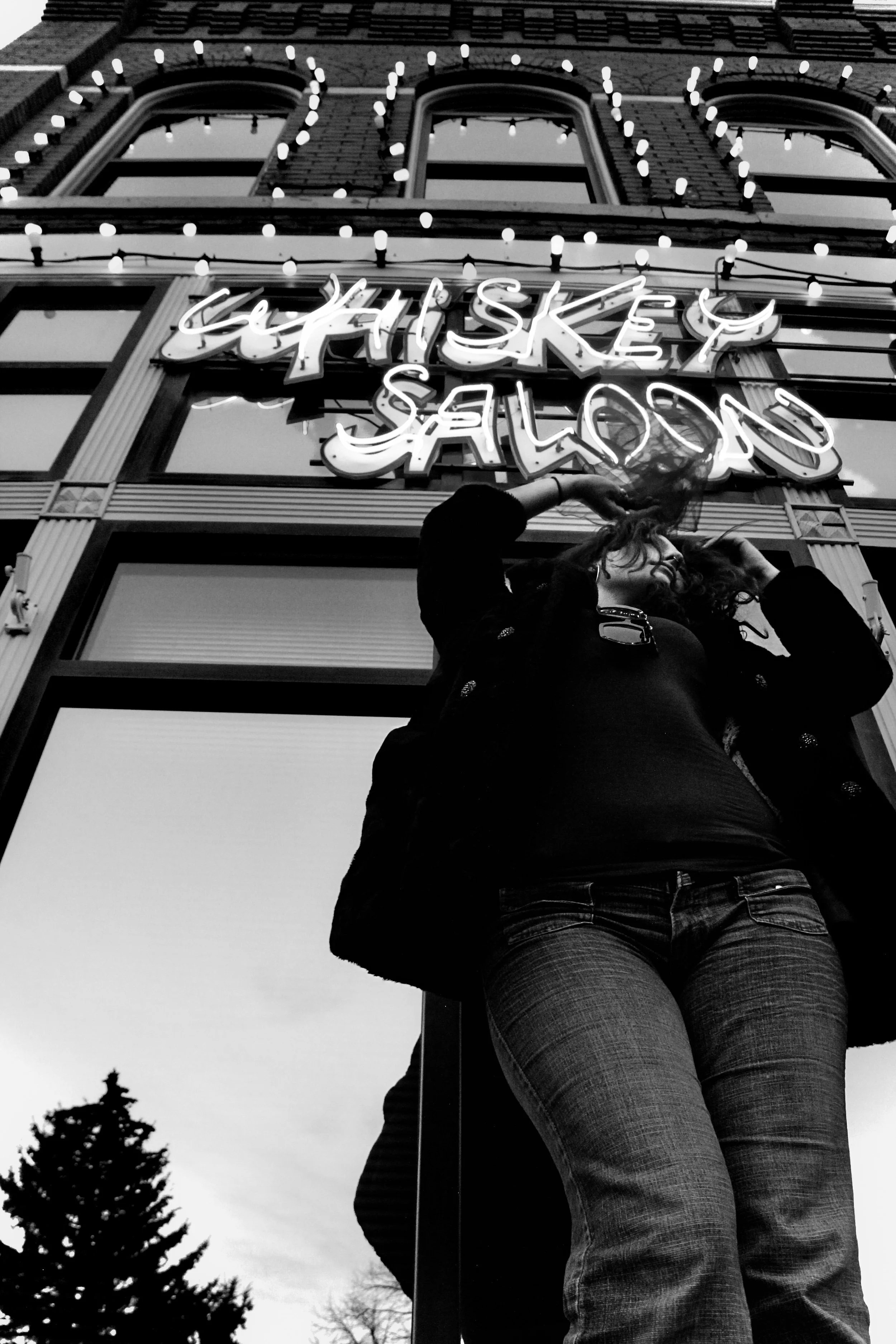 A woman is standing outdoors in front of a building with a neon sign that reads 'Whiskey Saloon,' adjusting her hair. The photo is taken from a low angle, and the sky is overcast.