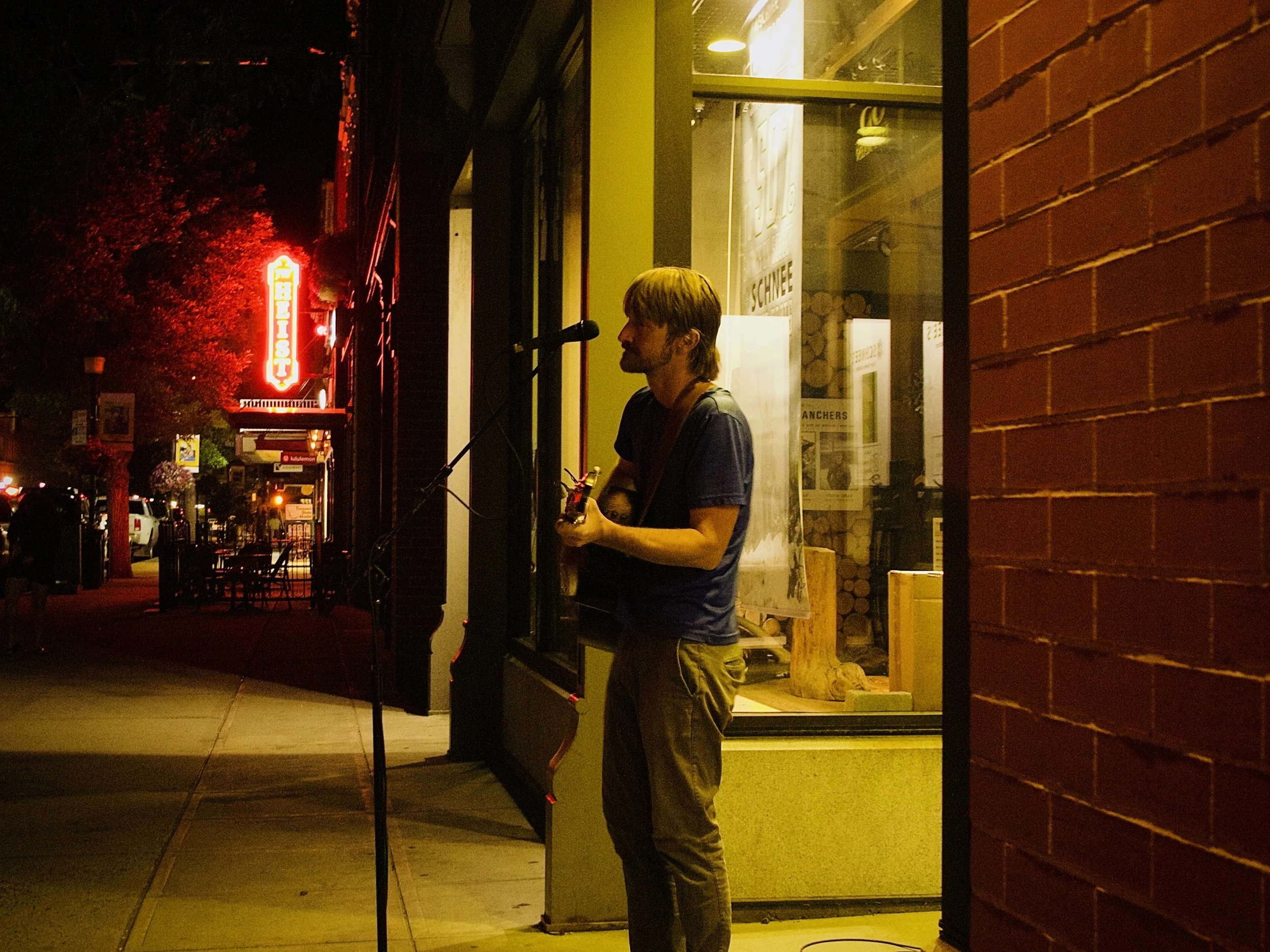 A musician with shoulder-length blond hair and a beard stands outside at night playing a guitar. He leans against a yellow wall, near a glass window with posters inside. A neon sign with the word 'BISTRO' glows in the background on a street lined wit