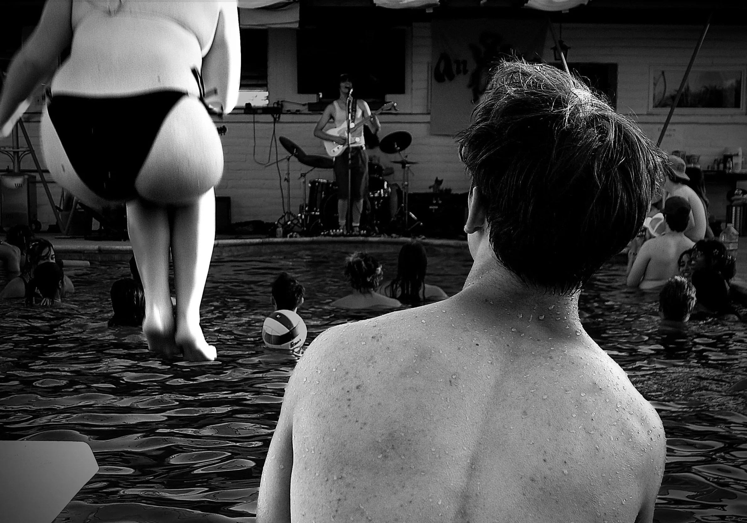 A woman with wet hair sitting in a swimming pool, observing a band performing on a stage outdoors. The band features a female musician playing an electric guitar and singing into a microphone with a drum set behind her. The scene includes other pool 