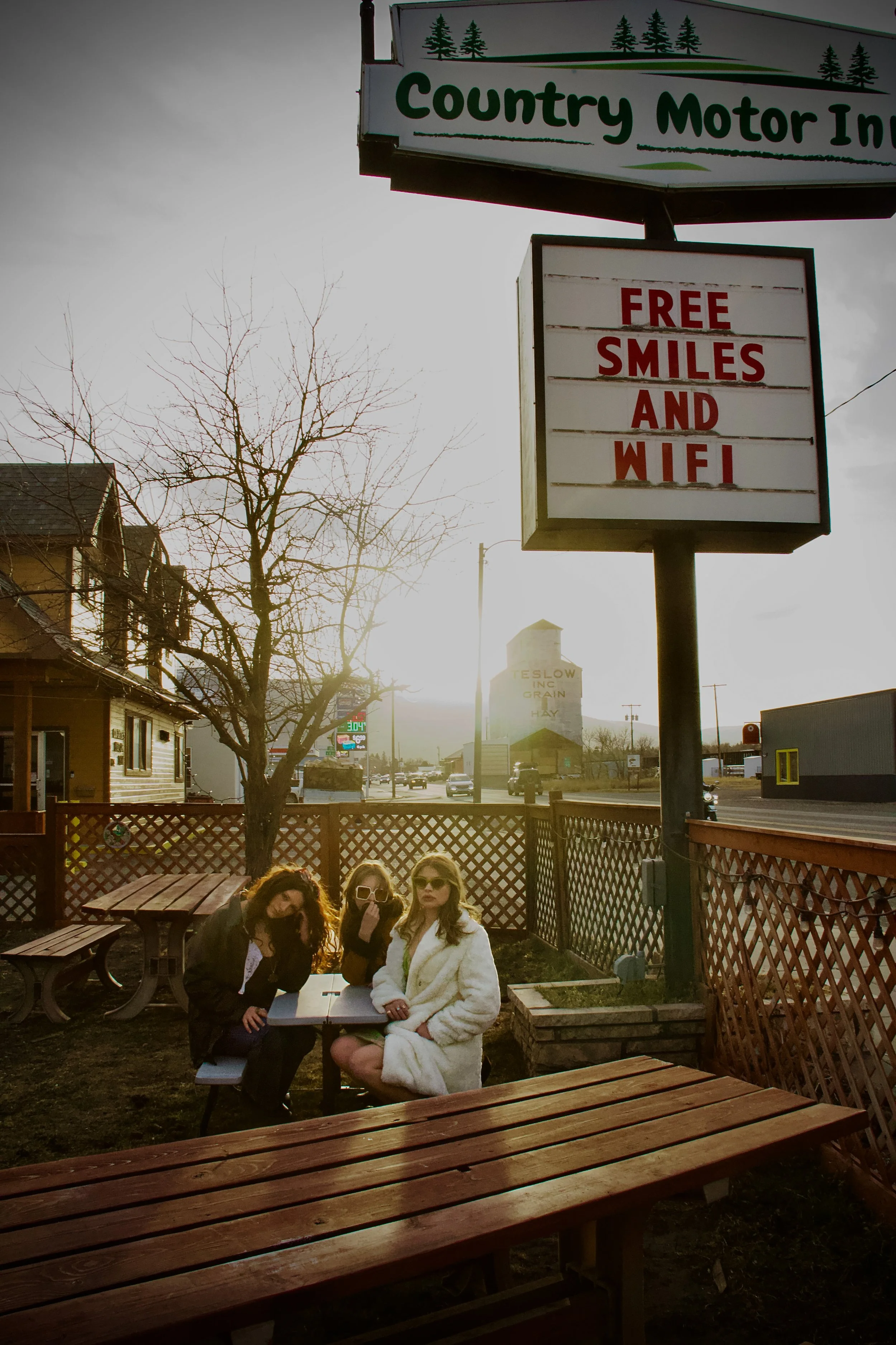 Three women sitting outside at a wooden picnic table beneath a sign for a country motel, which advertises free smiles and Wi-Fi. The women are wearing sunglasses and relaxed clothing, with one woman in a white robe. Trees and buildings are visible in