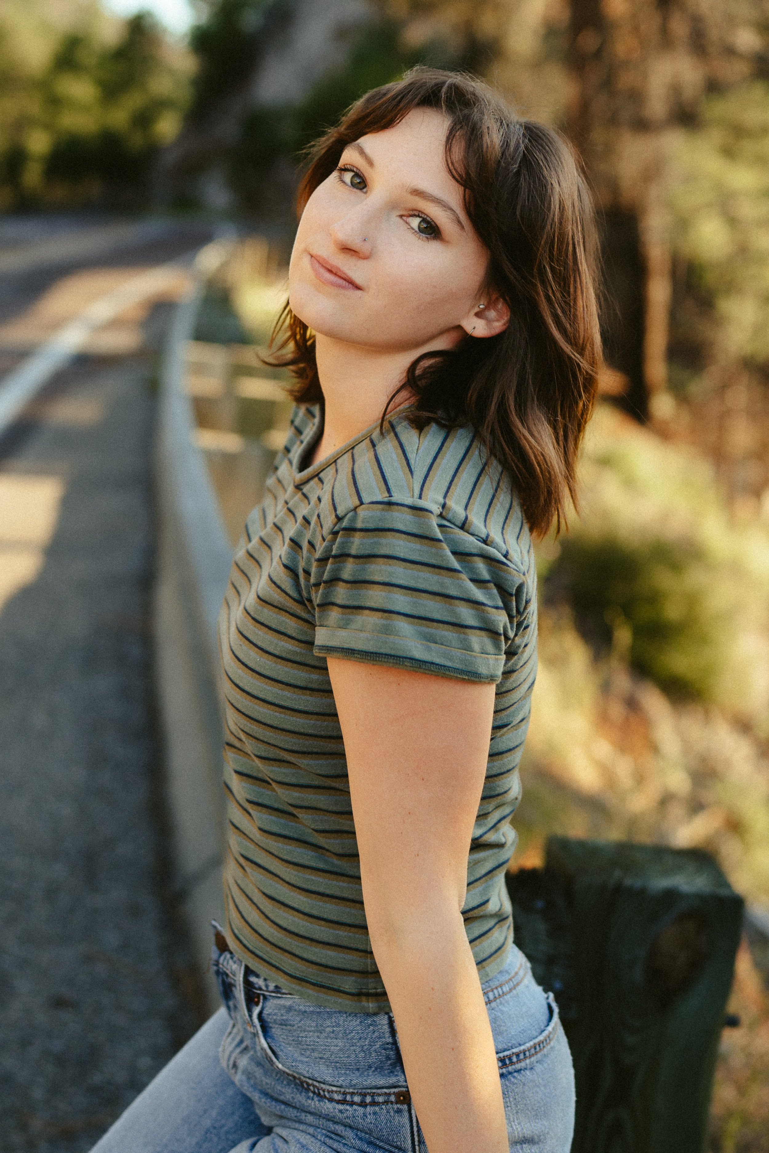 A young woman with shoulder-length brown hair and light skin leaning against a metal railing outdoors during daytime, wearing a striped t-shirt and jeans, with a background of trees and logs.