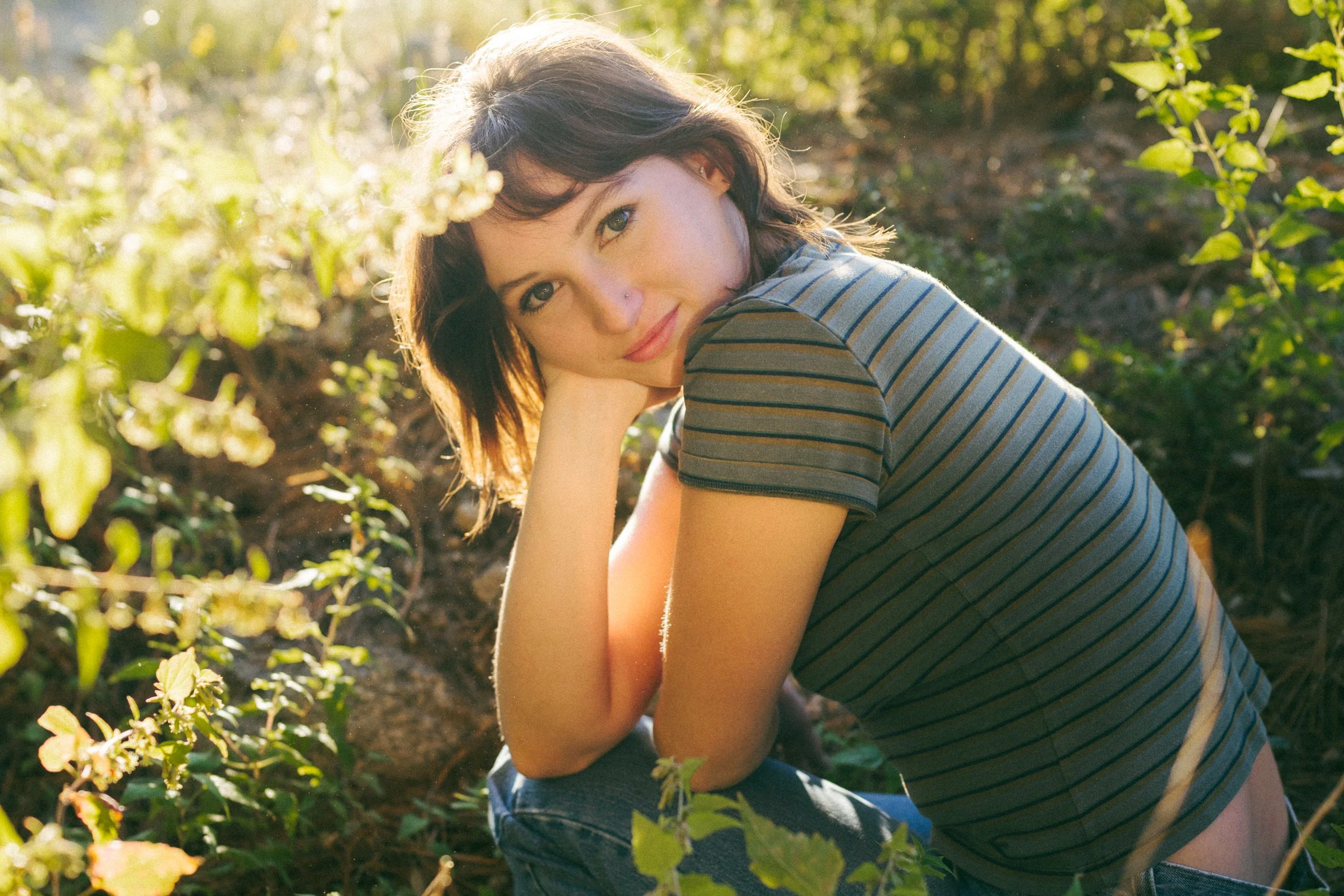 A young woman with short brown hair and a nose piercing sitting outdoors among green foliage, resting her chin on her hand, wearing a striped t-shirt and looking at the camera with a slight smile.