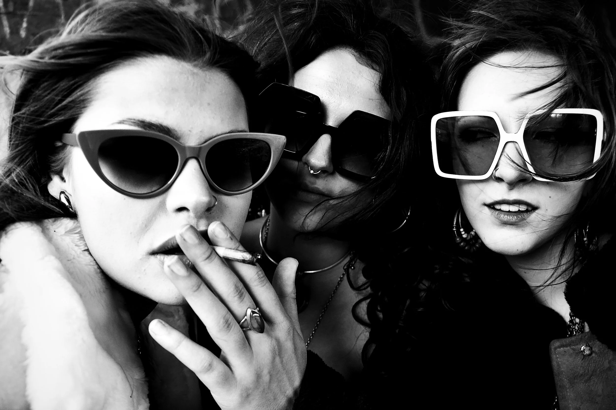 Three young women with dark hair and sunglasses, close-up portrait, black and white photo, they are lying down together and looking at the camera.