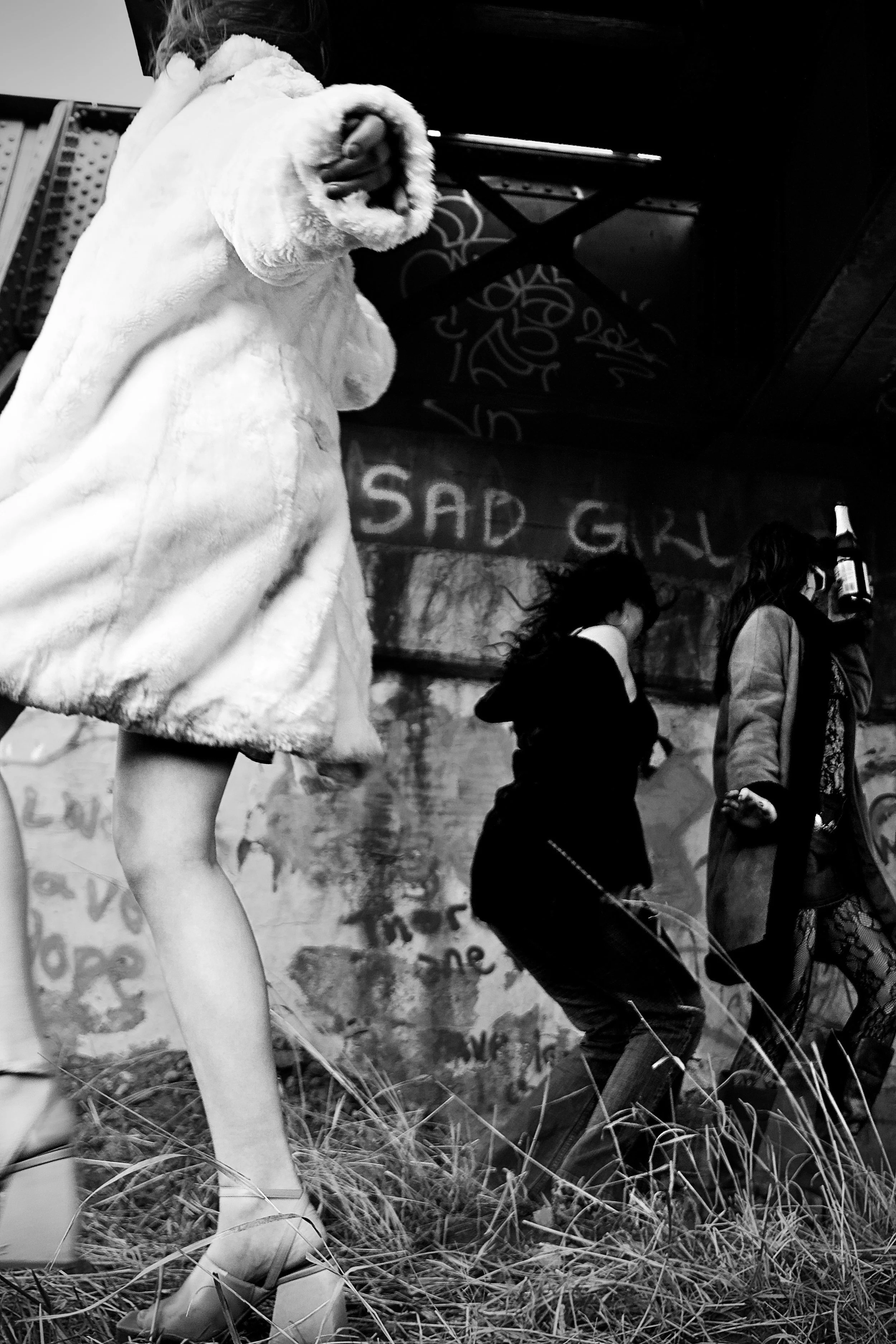 Black and white photo of a mannequin wearing a fluffy coat, with a focus on its legs. In the background, there are people, one of whom appears to be walking a dog. A sign reading 'SAD GIRL' is visible on the wall behind them.